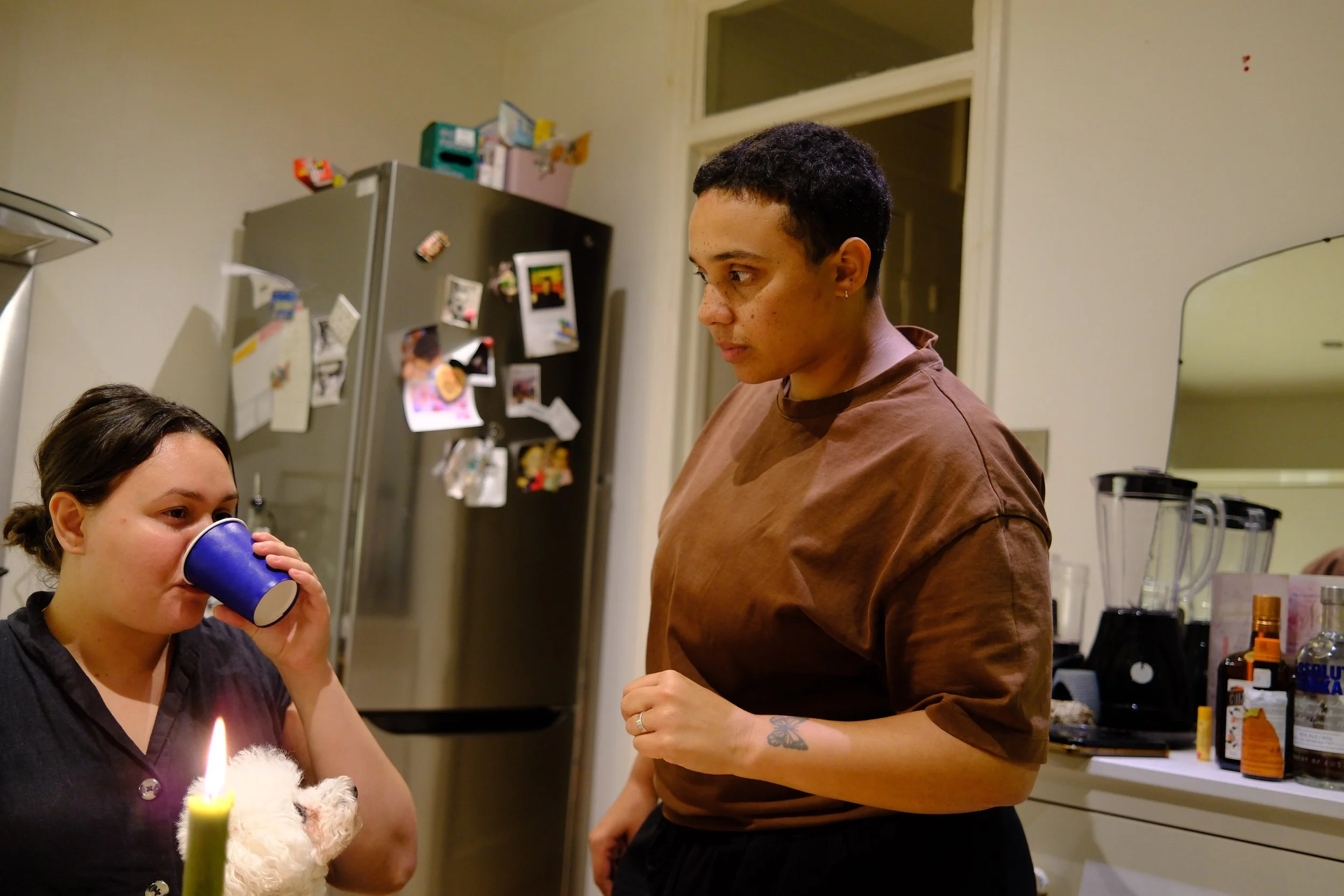 Two women in a kitchen, one drinking from a blue cup and holding a small white dog, while the other woman stands nearby looking at her.