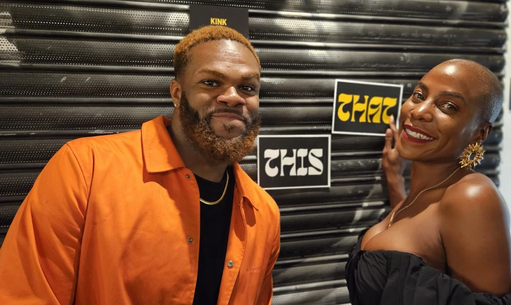 A man with blonde hair, a beard, and wearing an orange shirt standing next to a smiling woman with a shaved head, gold earrings, and a black strapless top. They are in front of a black backdrop with signs that say 'THIS' and 'THAT.'