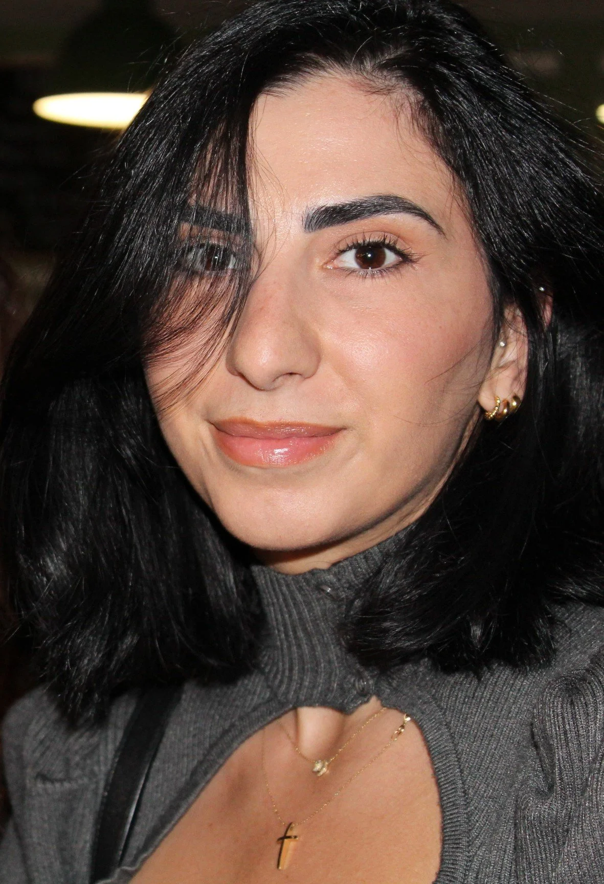 A woman with dark hair and brown eyes, wearing gold earrings and layered necklaces, smiling with minimal makeup in a grey top with a ruffled collar.