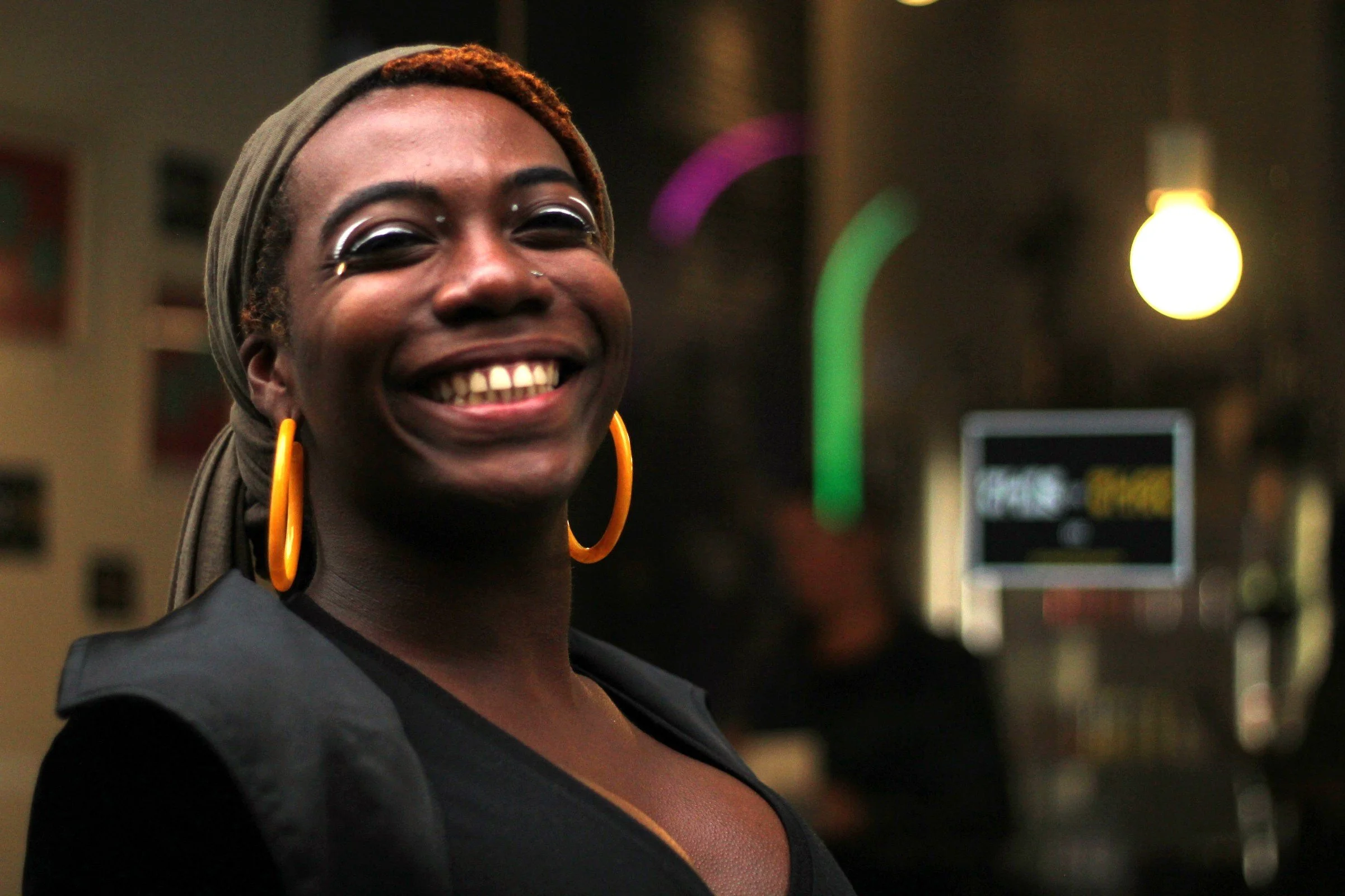 A smiling woman with dark skin, wearing large orange hoop earrings, a headscarf, and makeup with white eyeliner, standing indoors with colorful lights in the background.