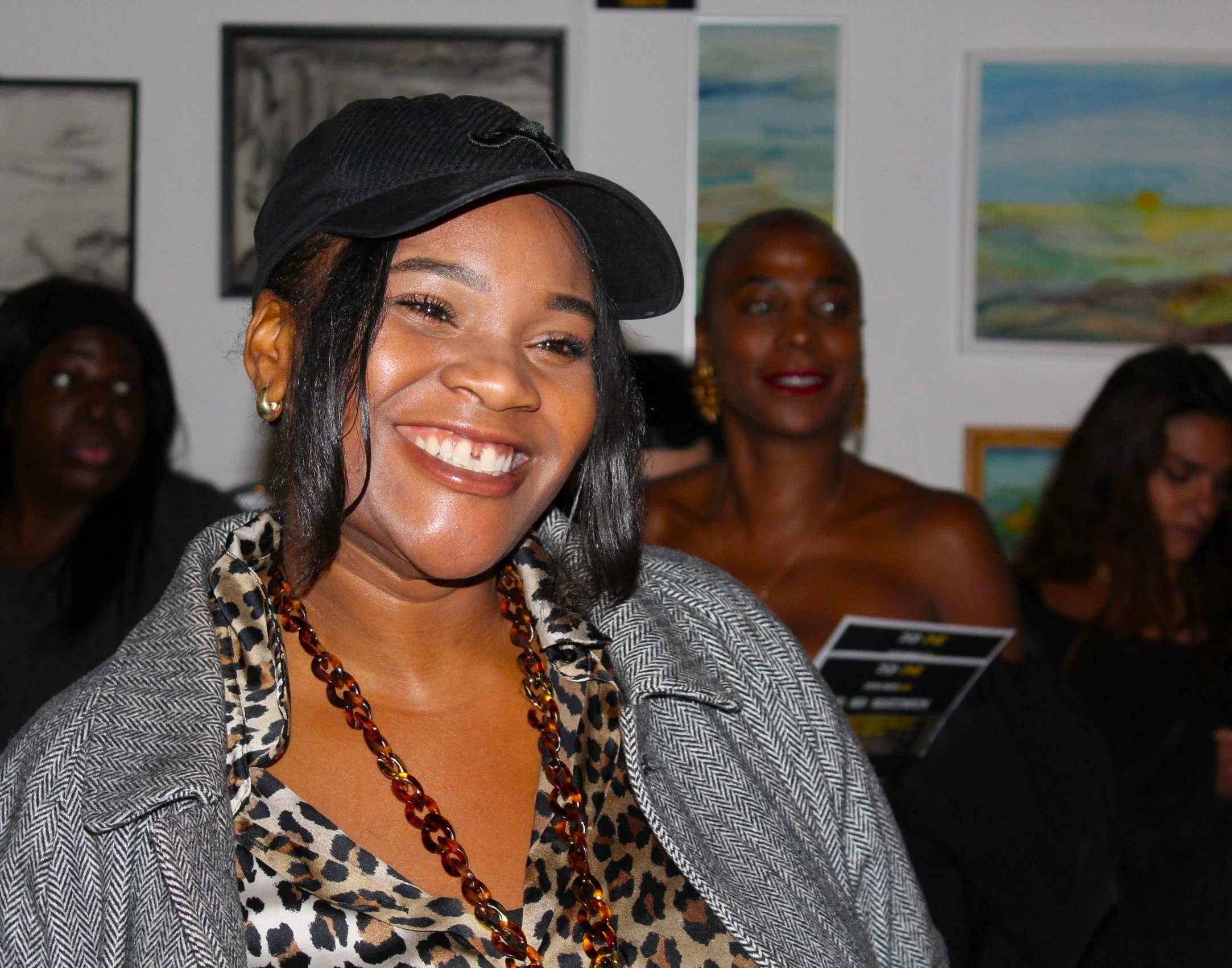 A group of women attending an event, with one woman smiling at the camera in the foreground, wearing a black cap, a leopard print top, a grey herringbone jacket, and a chunky amber necklace, with others in the background examining programs and engaging.