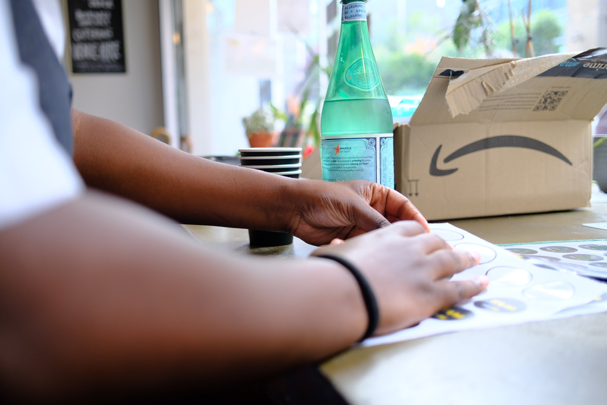 Person assembling stickers or labels on a table inside a cafe or shop, with a bottle of sparkling water, paper cups, a cardboard box with the Amazon logo, and some plants and windows in the background.