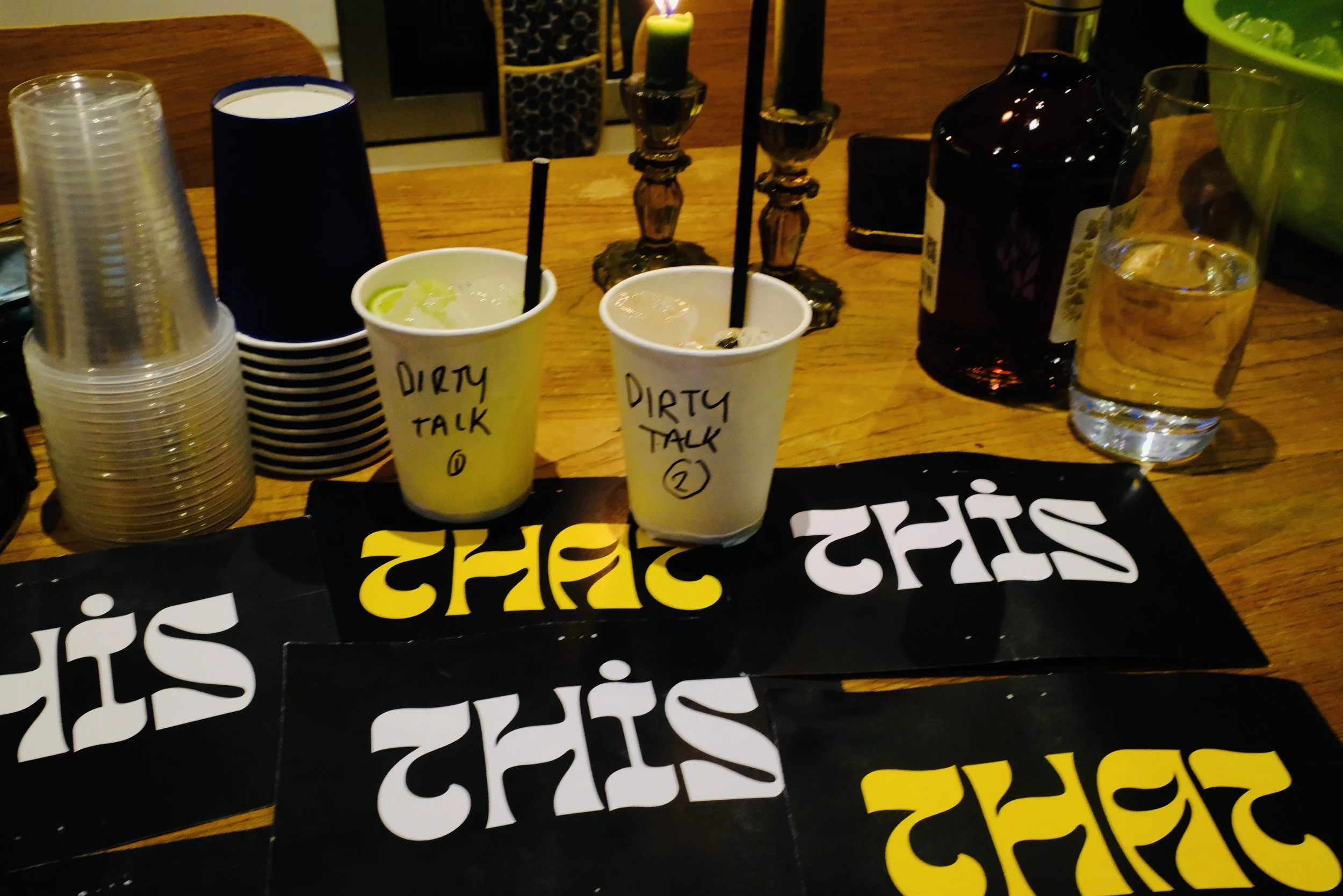 Two cups labeled 'Dirty Talk 1' and 'Dirty Talk 2' with drinks and black straws on a table, surrounded by plastic cups, a candle holder with candles, a bottle, and a glass of water.