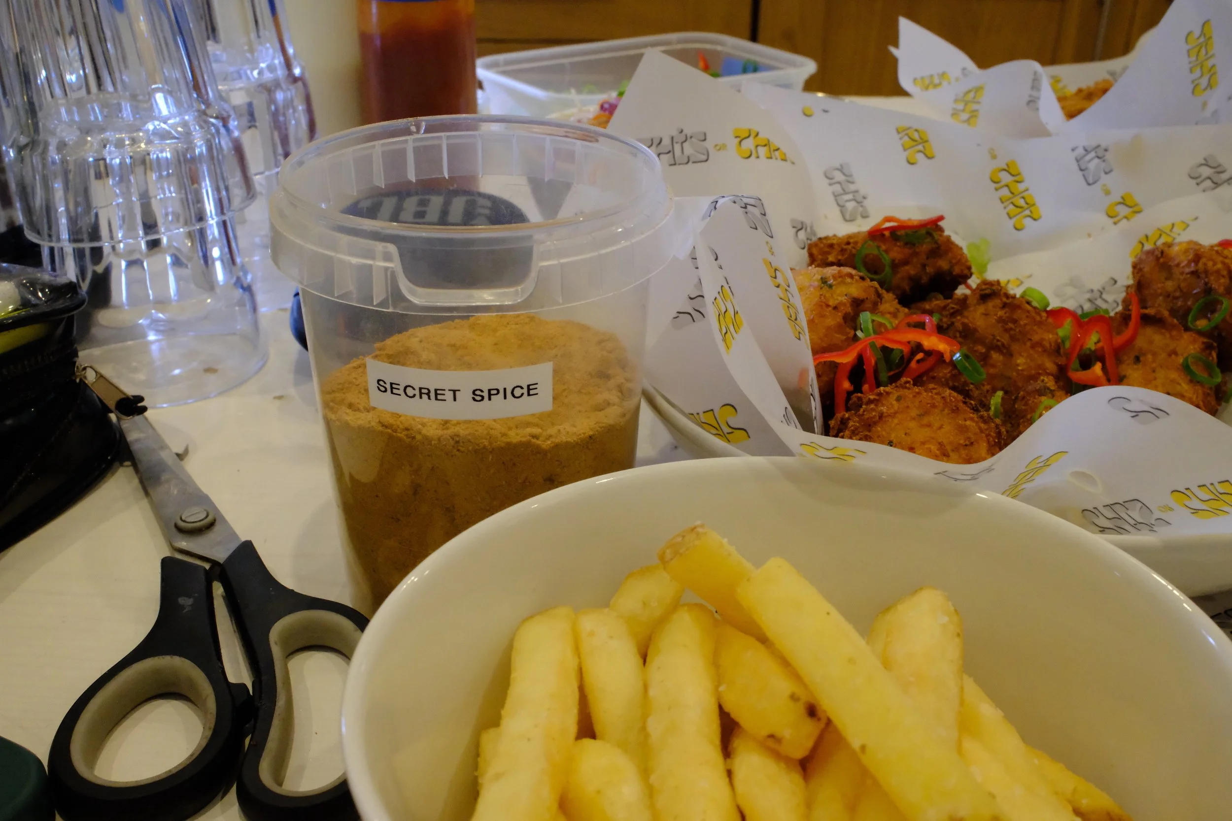 Basket of fried chicken pieces garnished with sliced green onions, a container of yellow spice labeled 'Secret Spice,' a bowl of French fries, and various kitchen utensils and condiments on a table.