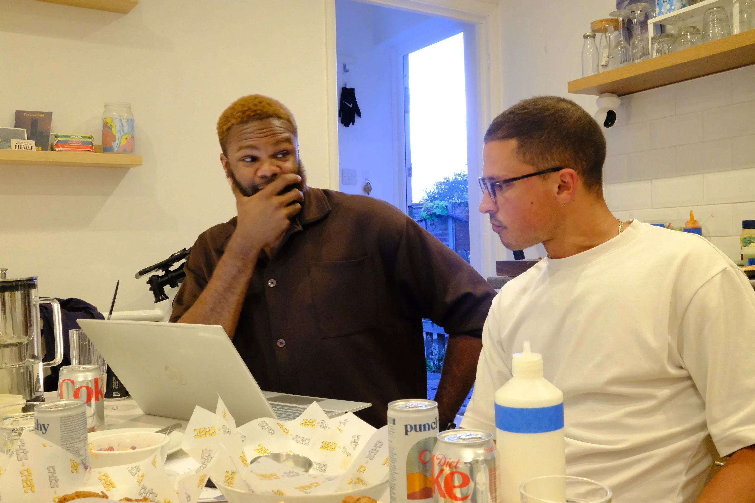 Two men sitting at a dinner table with food and drinks, one with glasses and a white shirt, the other with a brown shirt and a beard, having a conversation indoors.