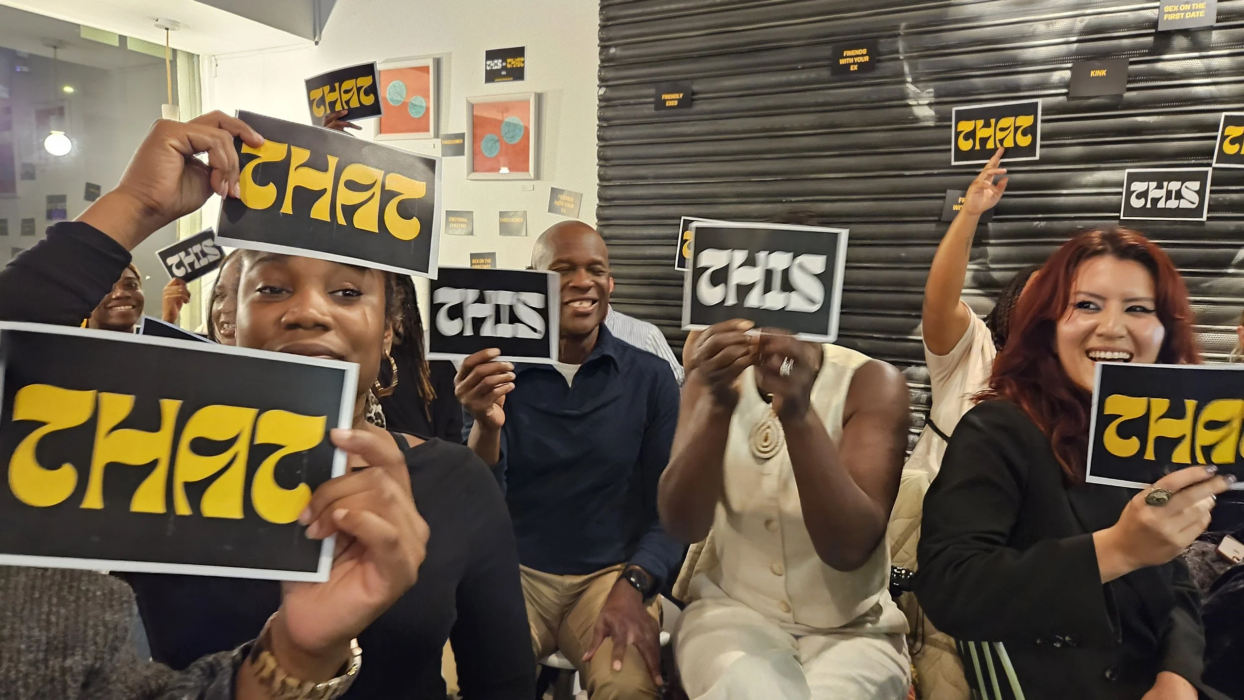 A group of people smiling and holding signboards with the word 'THAT' written in yellow on a black background, while sitting indoors.