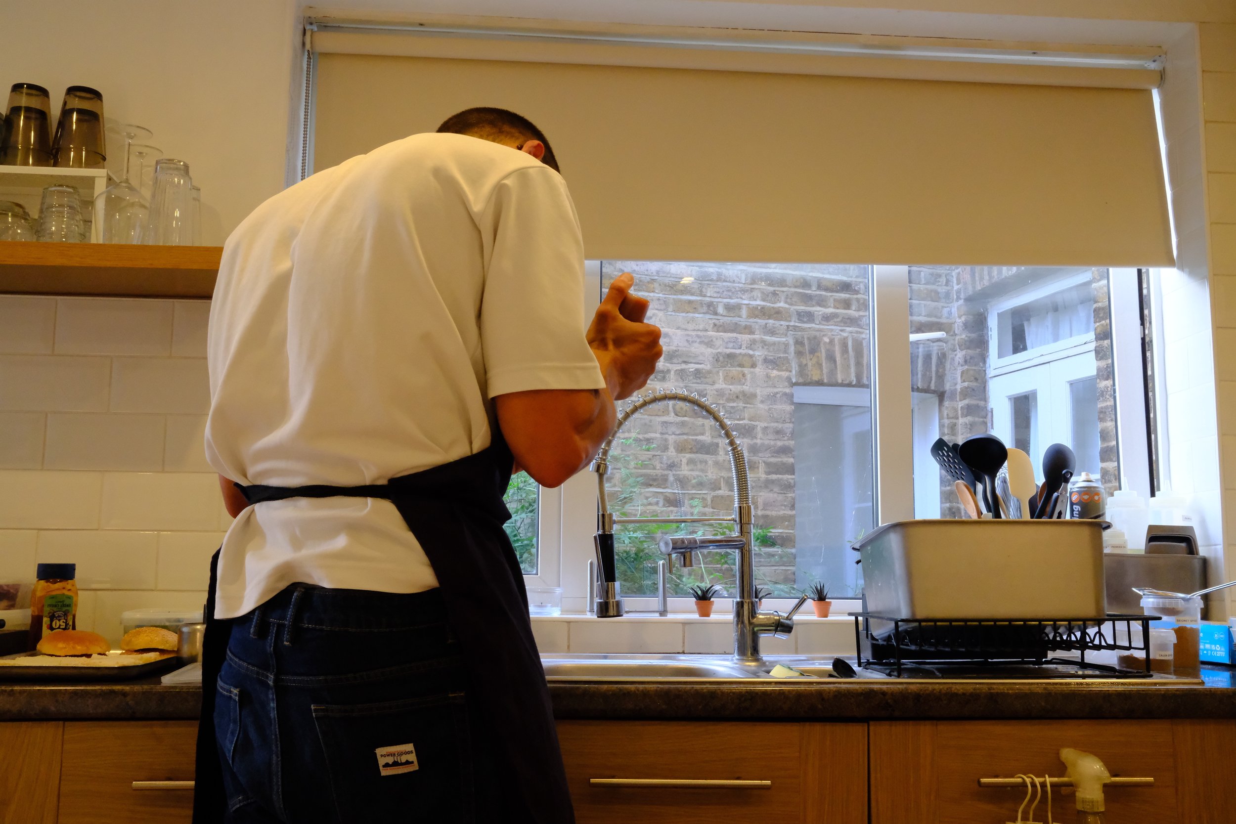 Person in a white shirt and dark apron standing at a kitchen sink, holding a knife near the window, with dishes and utensils organized around.
