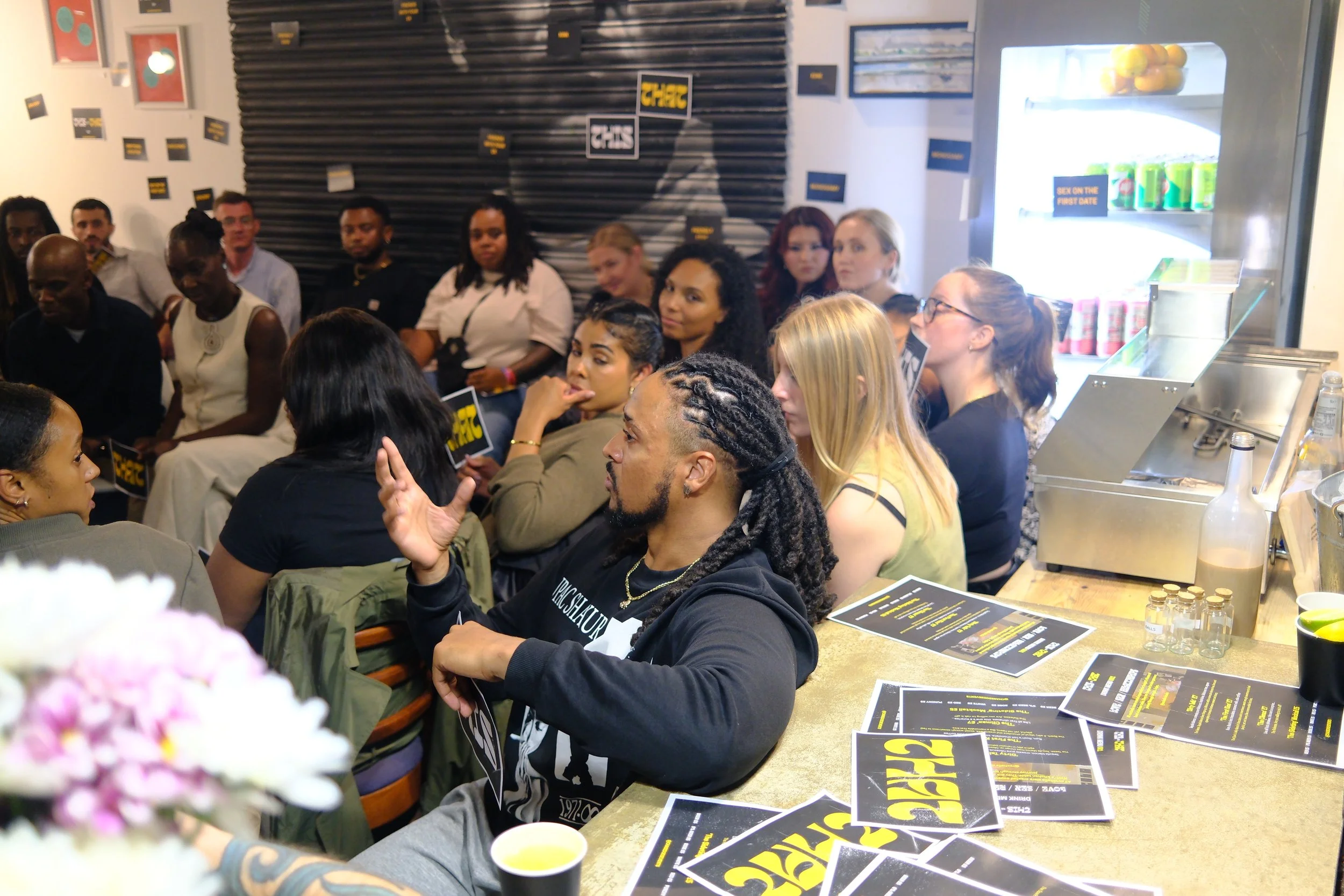 People gathered in a room attending a discussion or presentation, with some holding signs and papers, and a man speaking in the foreground.