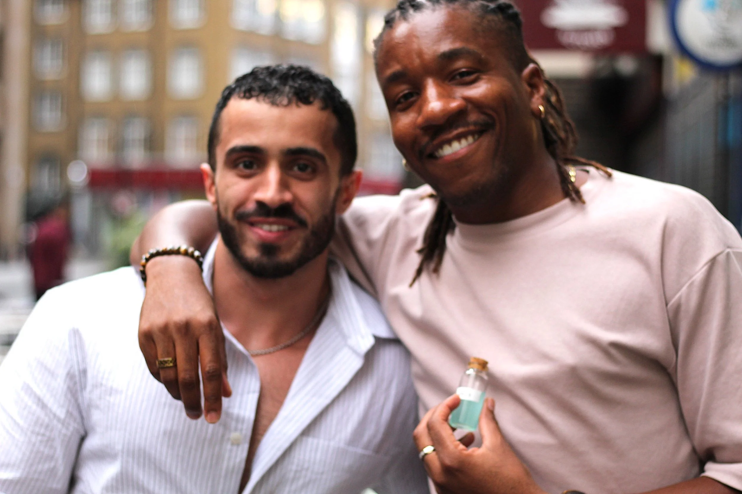 Two smiling men standing outdoors with arm around each other, one holding a small bottle of sanitizer. In the background, city buildings are blurred.