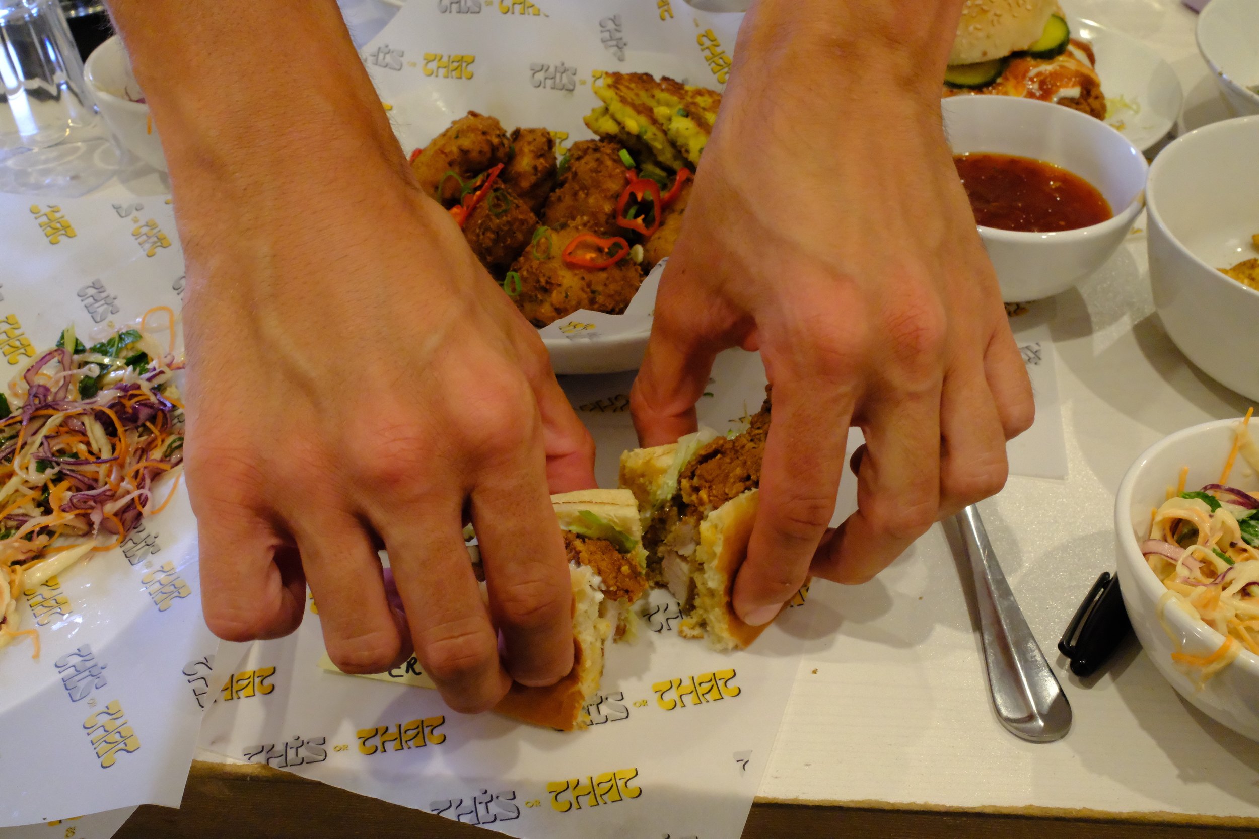 Person cutting a sandwich filled with fried chicken, lettuce, and sauce at a table with various side dishes and dipping sauces.