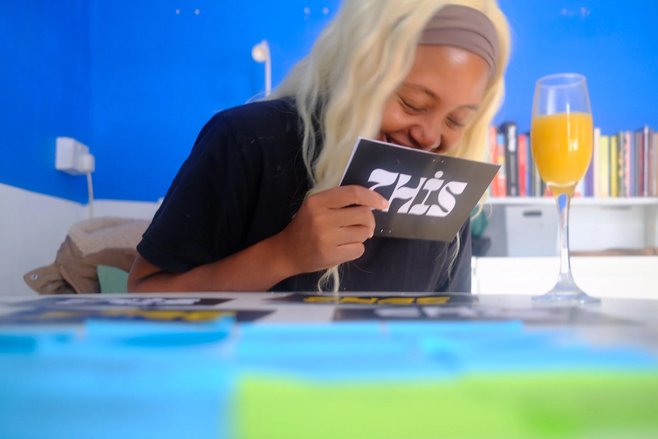 Woman with blonde hair and a gray headband smiling while looking at a black and white photo or card on a table. There is a glass of orange juice and a bookshelf in the background, with a blue wall behind her.