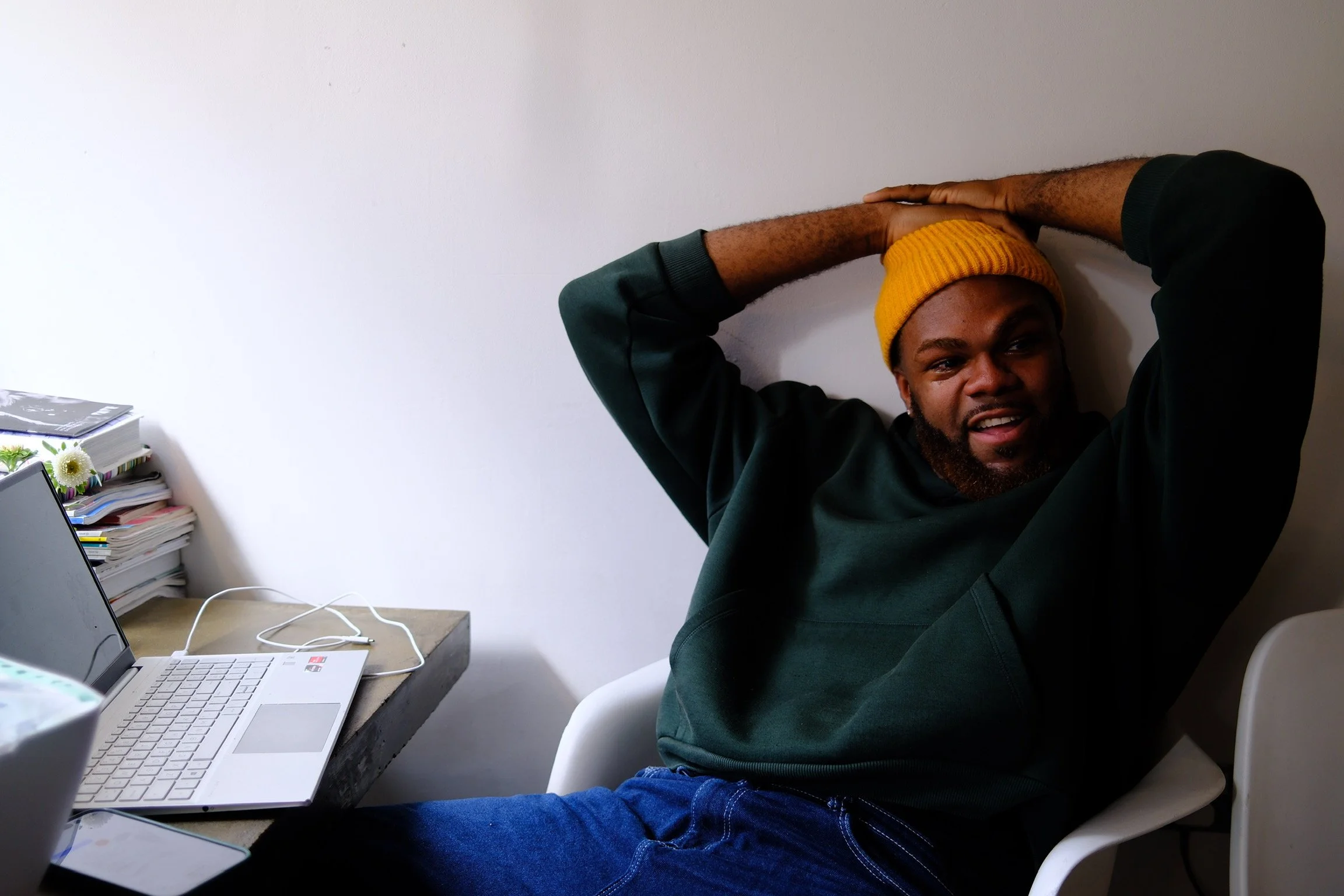 A man wearing a yellow beanie and a dark green sweatshirt sits in a white chair, leaning back with his hands on his head, smiling at the camera. He is beside a desk with a laptop and a stack of papers.
