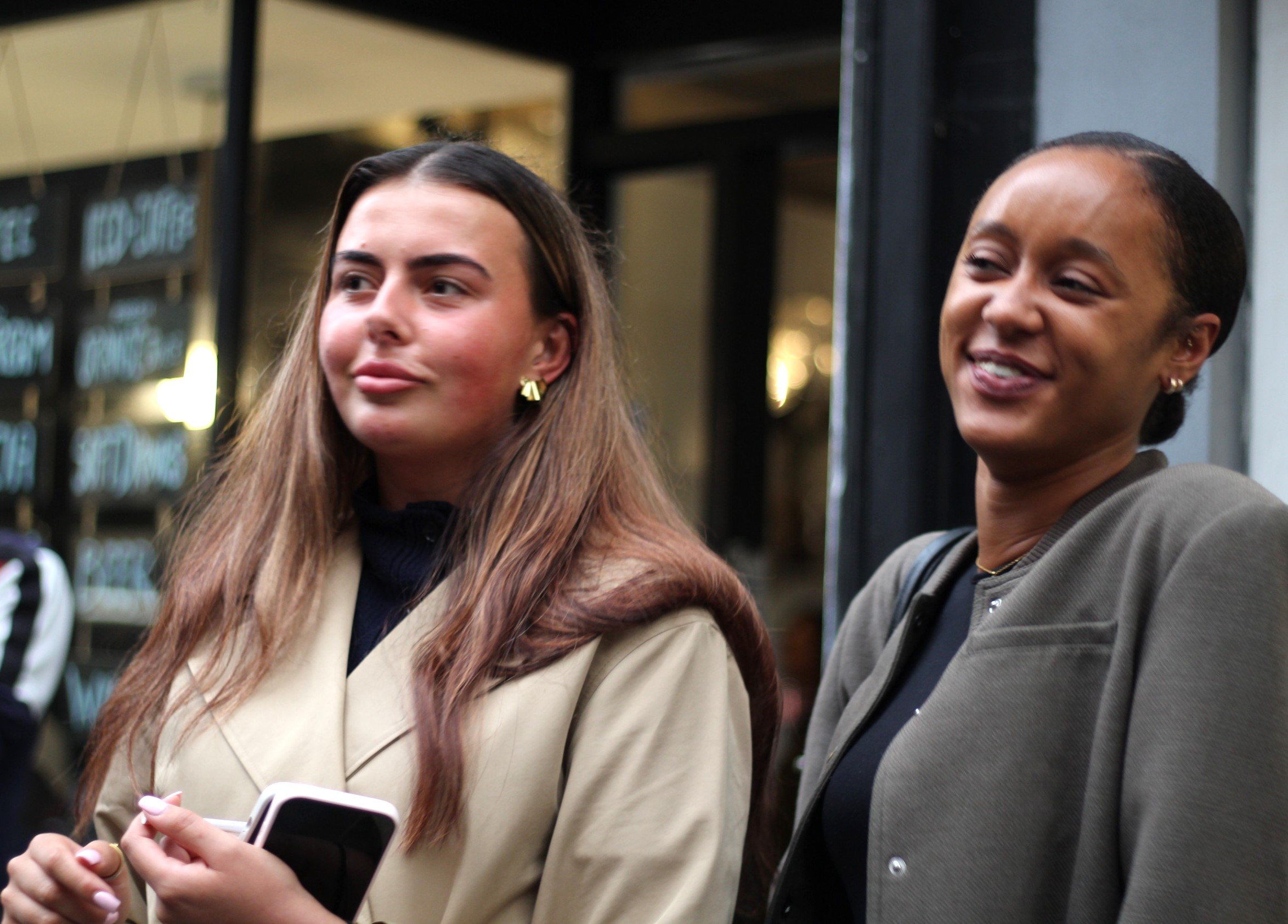 Two women standing outdoors, one holding a smartphone, smiling and looking to the side, background showing a storefront with illuminated signs.