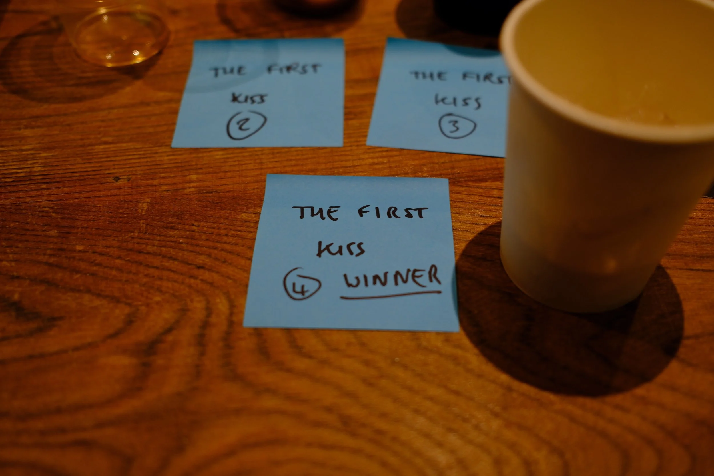 Three blue sticky notes on a wooden table with handwritten message 'The first kiss' and numbered circles 2, 3, and 4. Partially visible cup of coffee or tea is on the right side.
