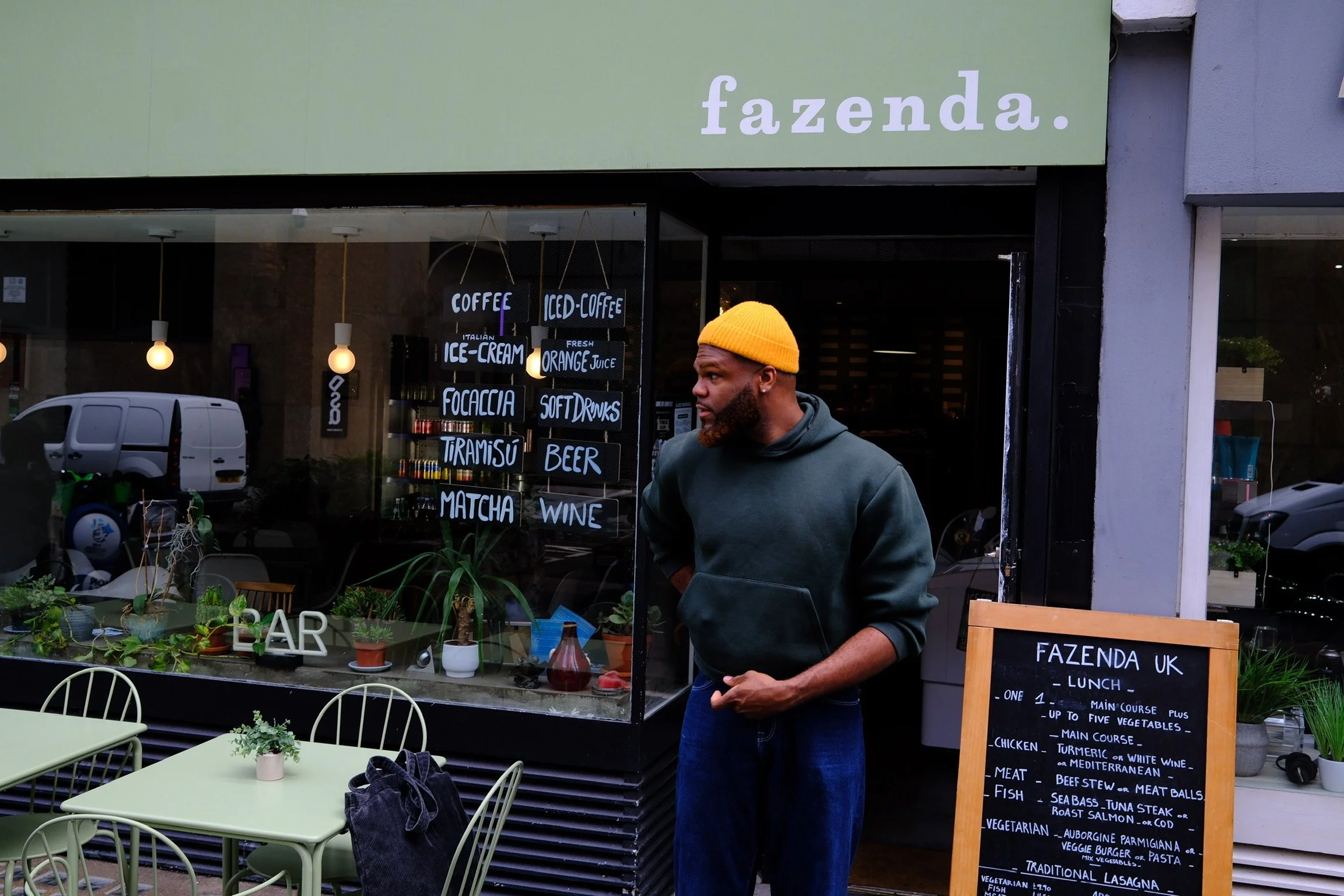 A man with a beard wearing a yellow beanie and green hoodie stands outside a café with a black chalkboard menu. The café's large window displays handwritten signs for coffee, ice cream, soft drinks, beer, wine, and fresh orange juice. Outdoor seating with light green tables and chairs is visible in the foreground.
