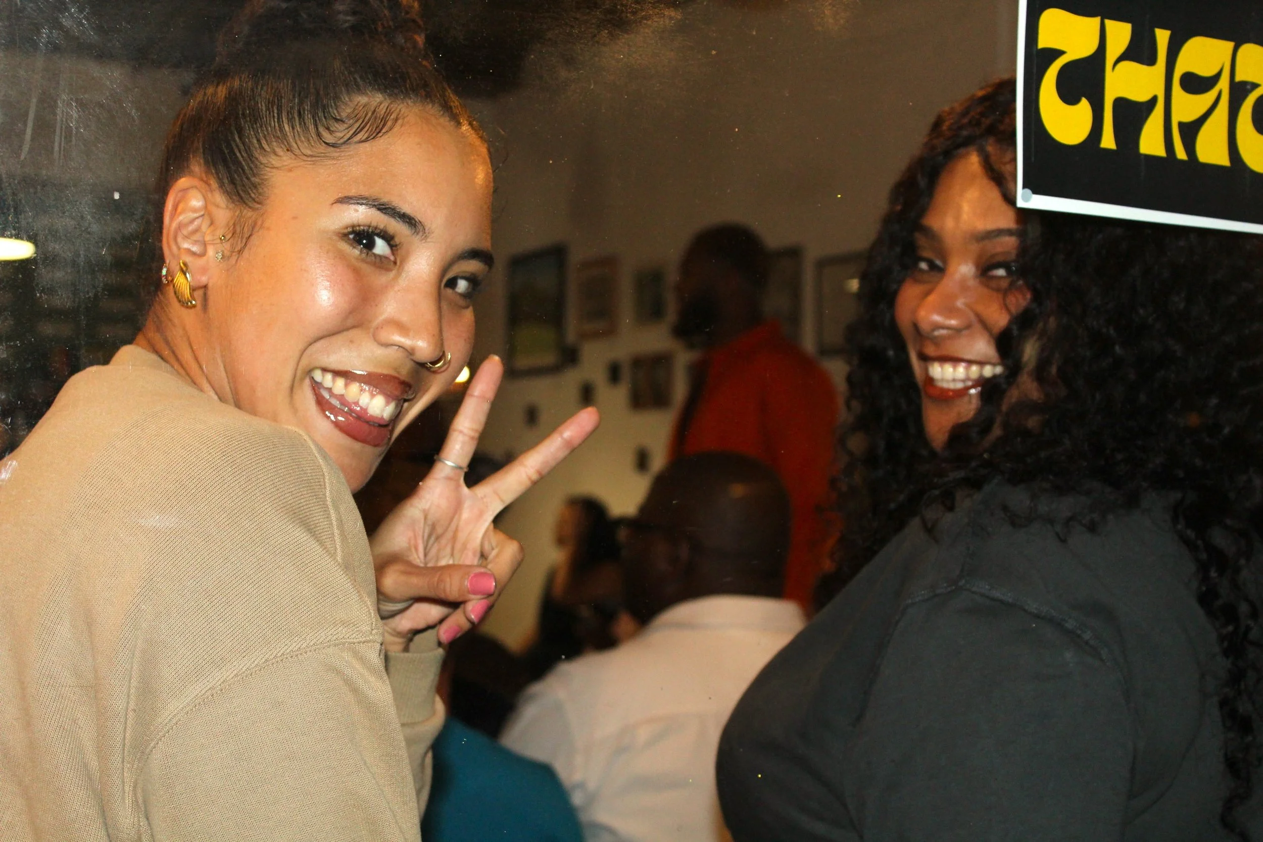Two women smiling and posing for a photo, one making a peace sign. They are in an indoor setting with other people in the background.