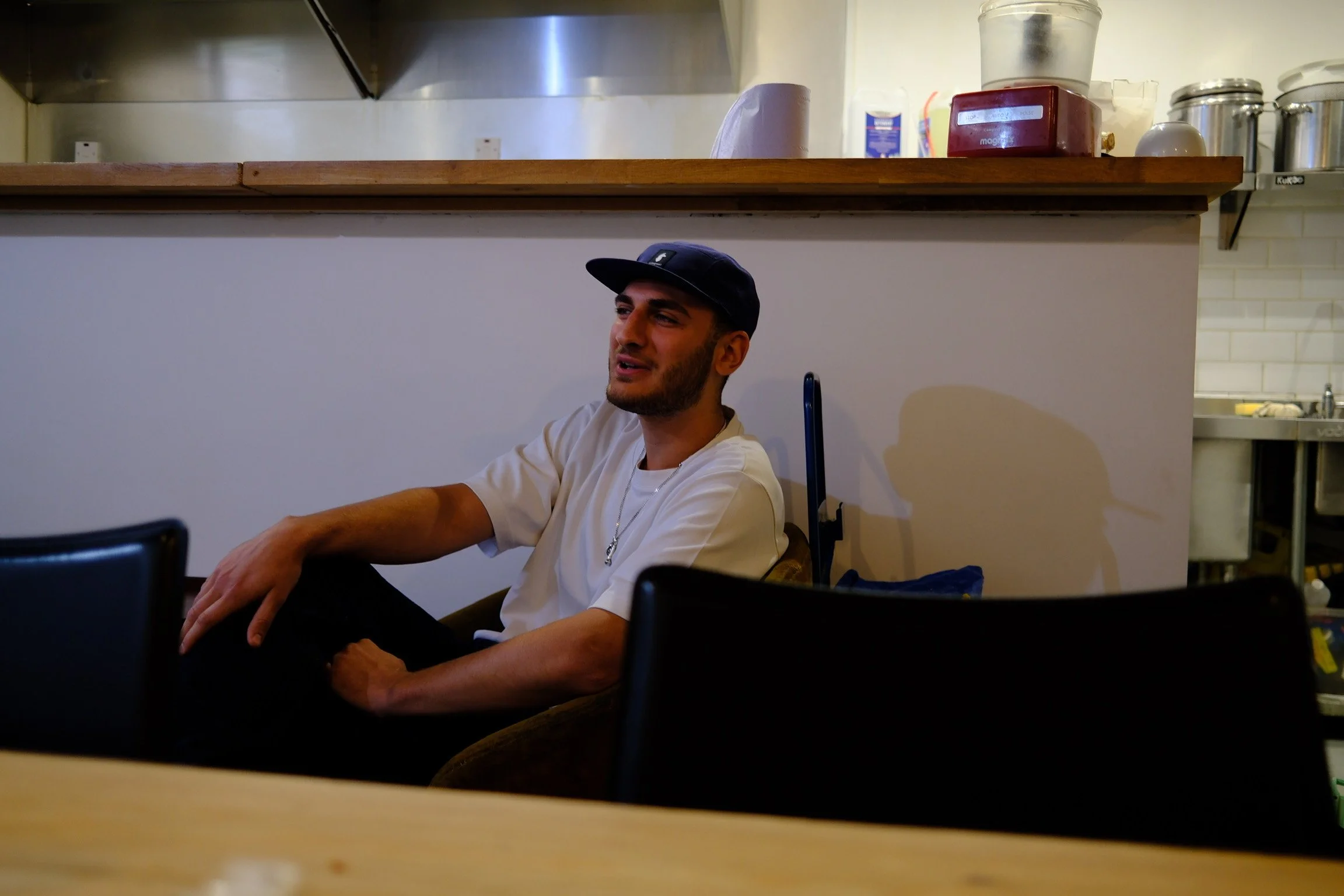 A young man with a beard, wearing a white t-shirt, a necklace, and a black baseball cap, sitting and leaning back in a chair in a café or restaurant, with a slight smile.