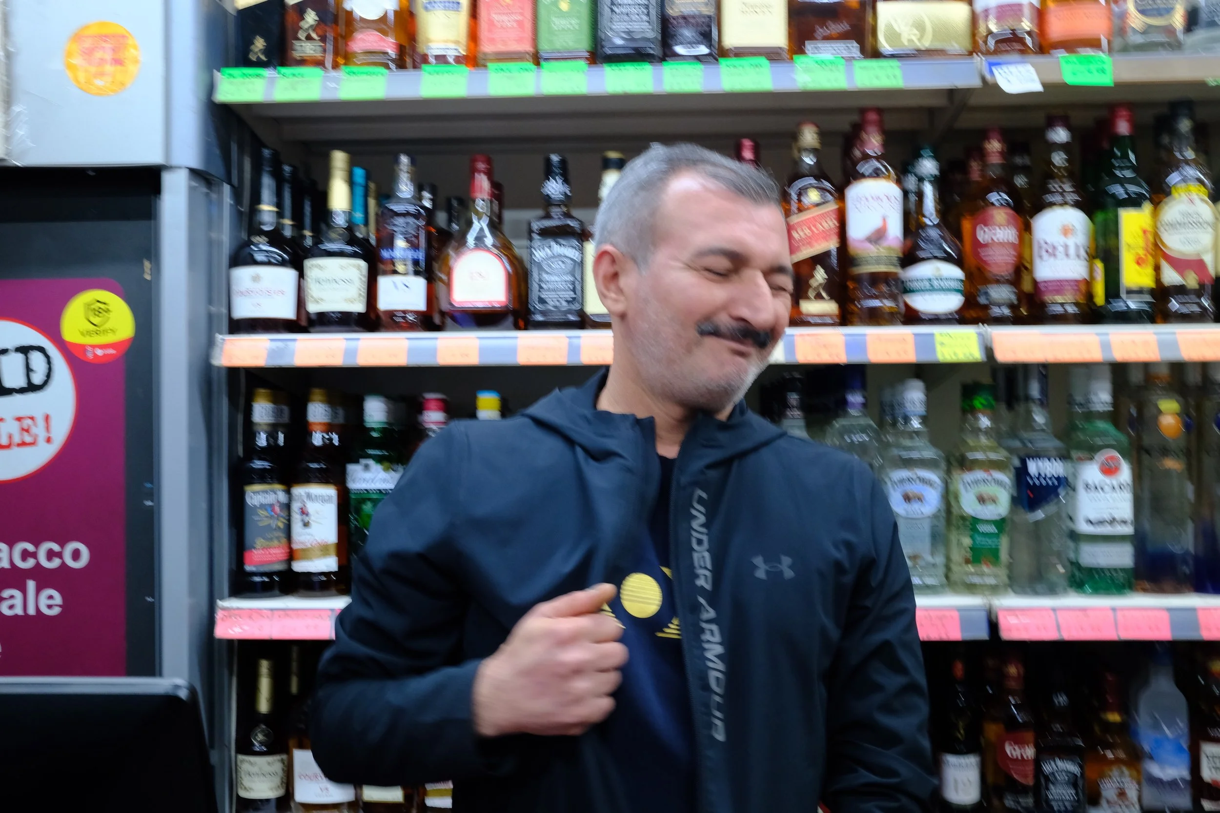 A man with a gray mustache and short gray hair standing in front of shelves of various bottles of alcohol at a store. He is wearing a black jacket and a dark shirt with a yellow and white logo on it.