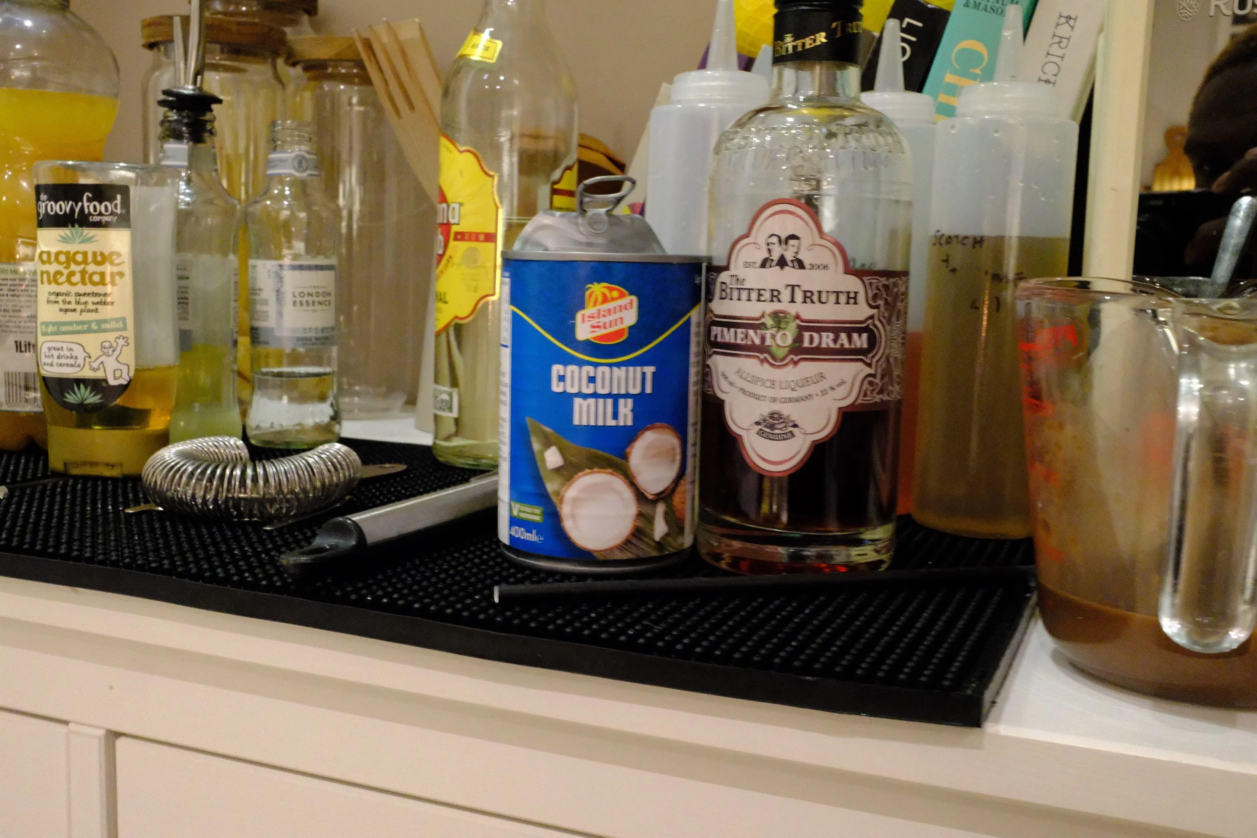 Bar counter with various liquor bottles, a can of coconut milk, a jar of agave nectar, some squeeze bottles, and a glass pitcher with a dark liquid and a metal spoon inside.