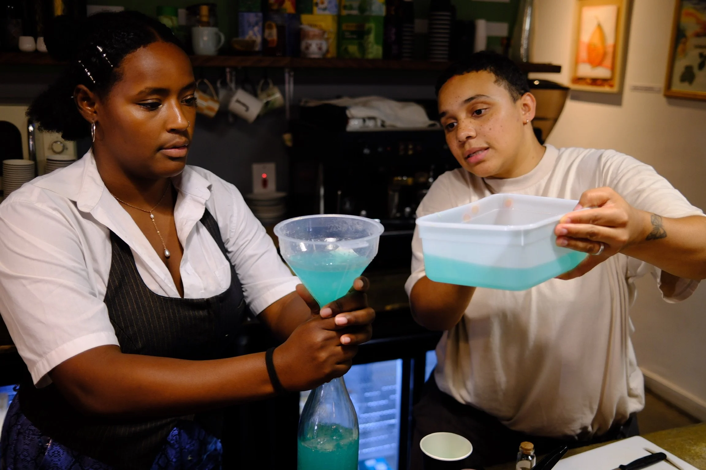 Two women are working together in a kitchen, pouring a blue liquid from a funnel into a rectangular container. The woman on the left is holding the funnel, and the woman on the right is holding the container. The background includes kitchen shelves with cups, jars, and framed pictures.