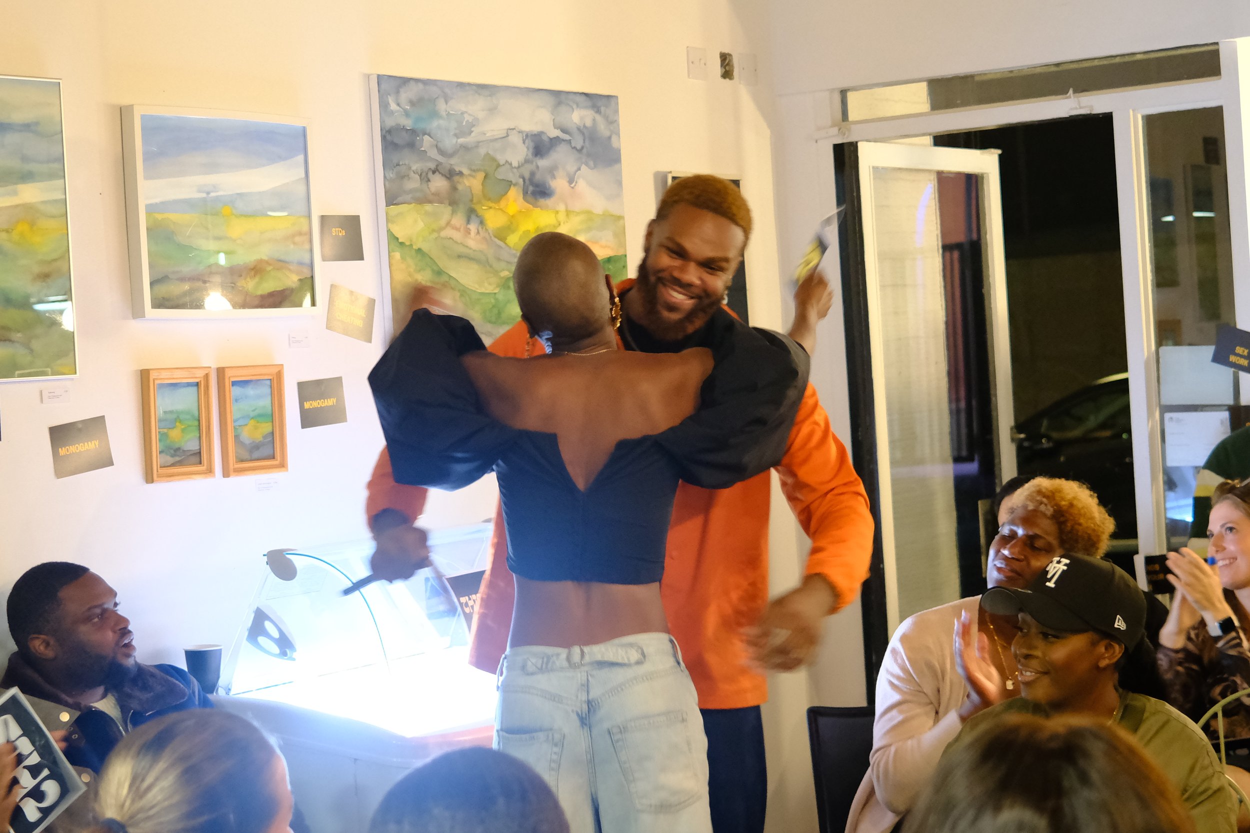 Two people hugging, smiling, in a lively art gallery. Several viewed artworks and audience members watching with smiles.