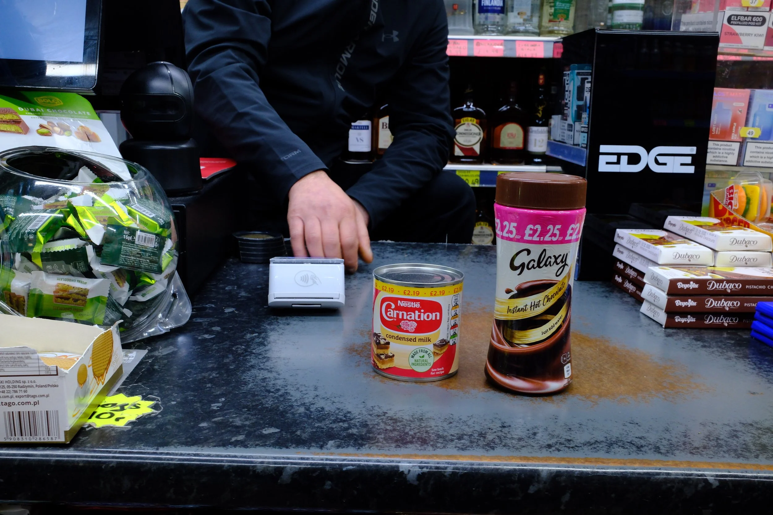 Counter at a grocery store checkout with a person in black clothing, two cans of condensed milk and hot chocolate mix, and various snacks and items around.