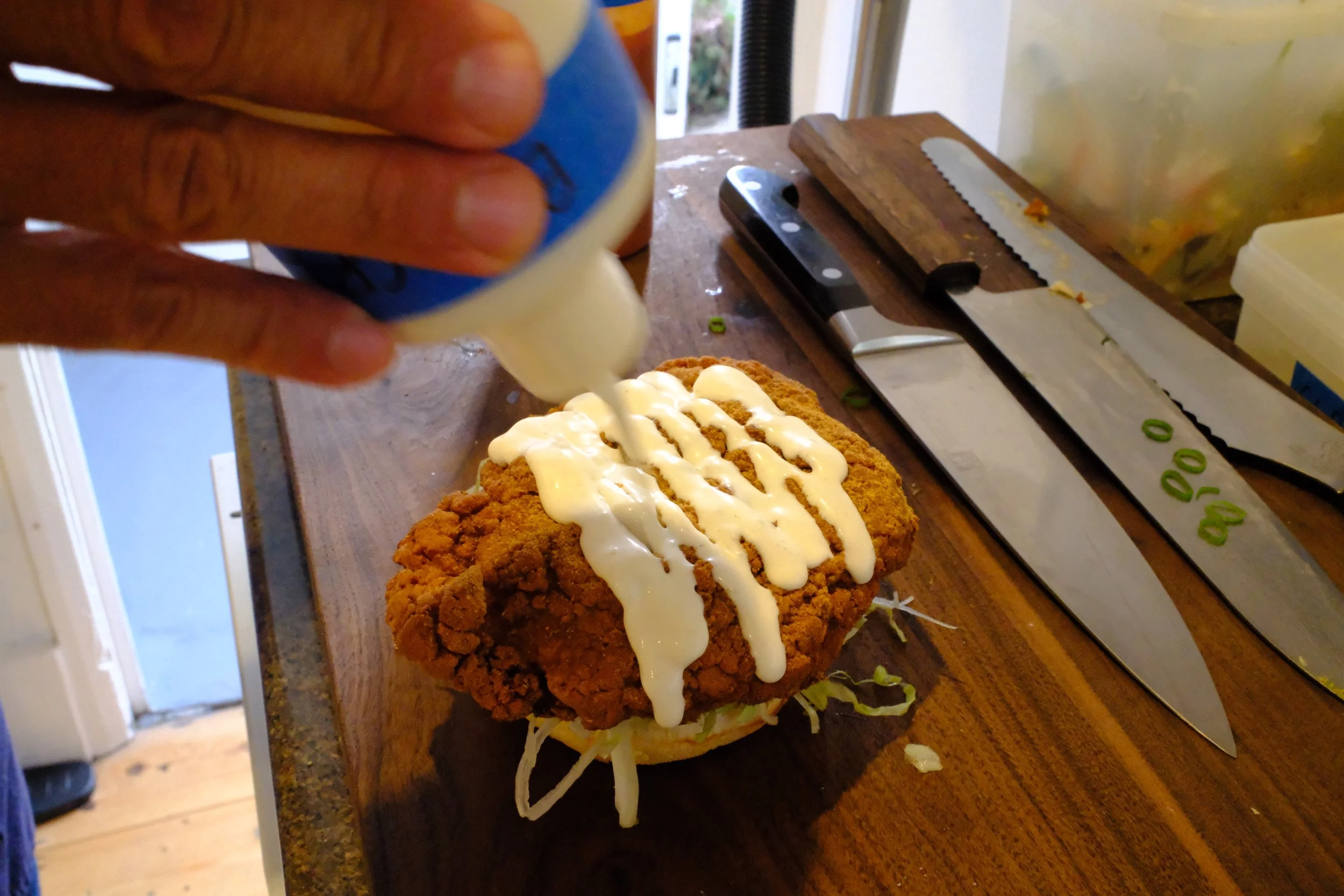 A fried chicken sandwich being topped with mayonnaise by a person in a kitchen. The sandwich is on a wooden countertop with a knife, a cleaver, and sliced green onions nearby.