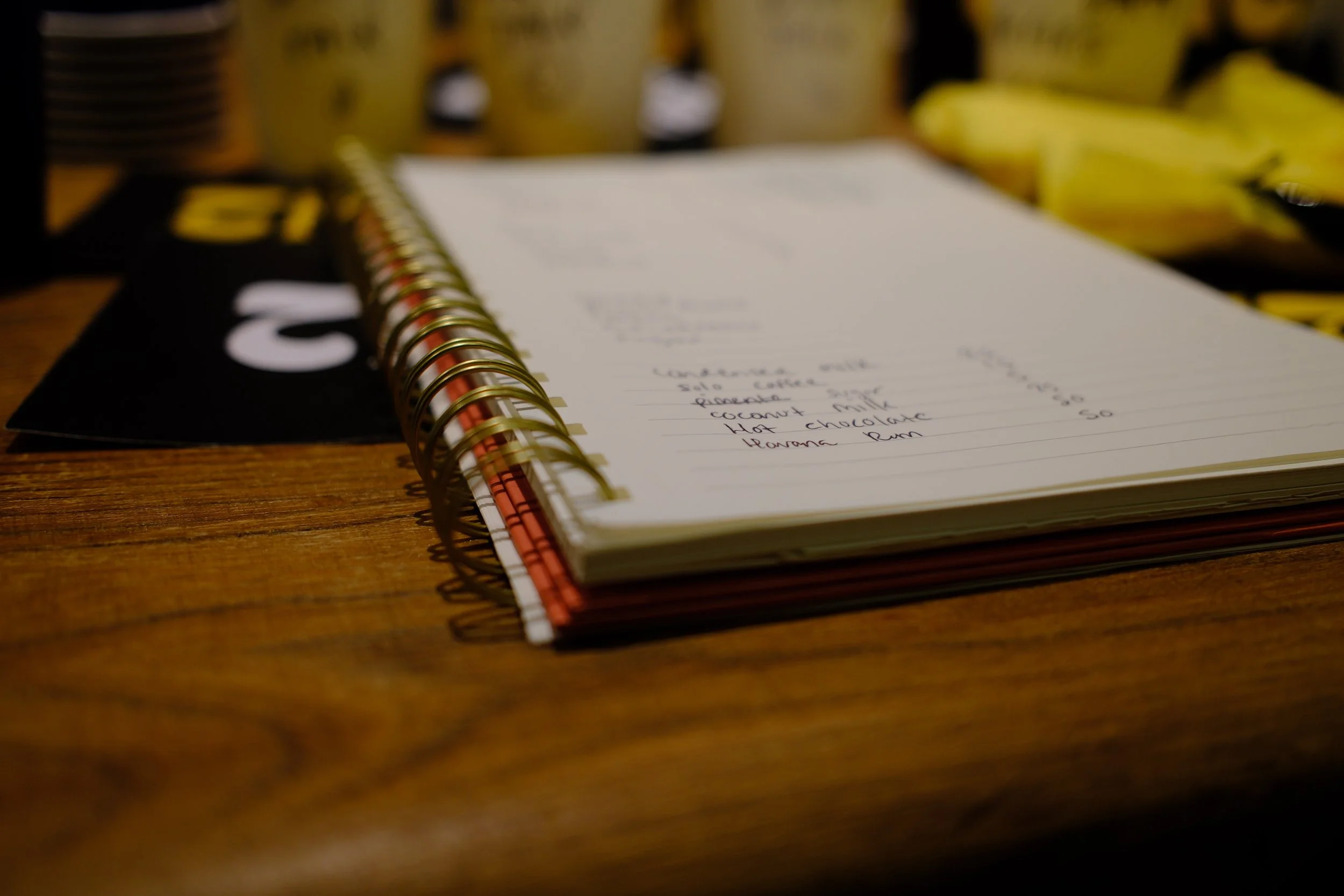 Open spiral-bound notebook resting on a wooden table, with a blurred background of bottles and bananas.