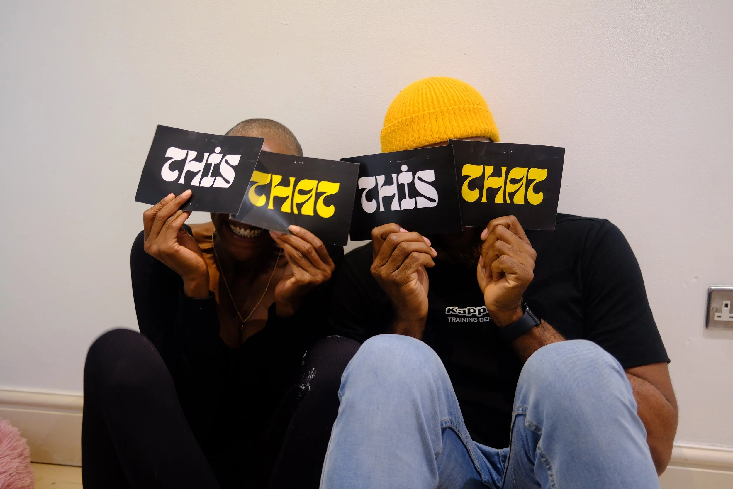 Two people sitting on the floor holding signs in front of their faces. The signs spell "THAT" in different fonts and colors. White person on the left with short hair and dark skin; person on the right wearing a yellow beanie and a black t-shirt.