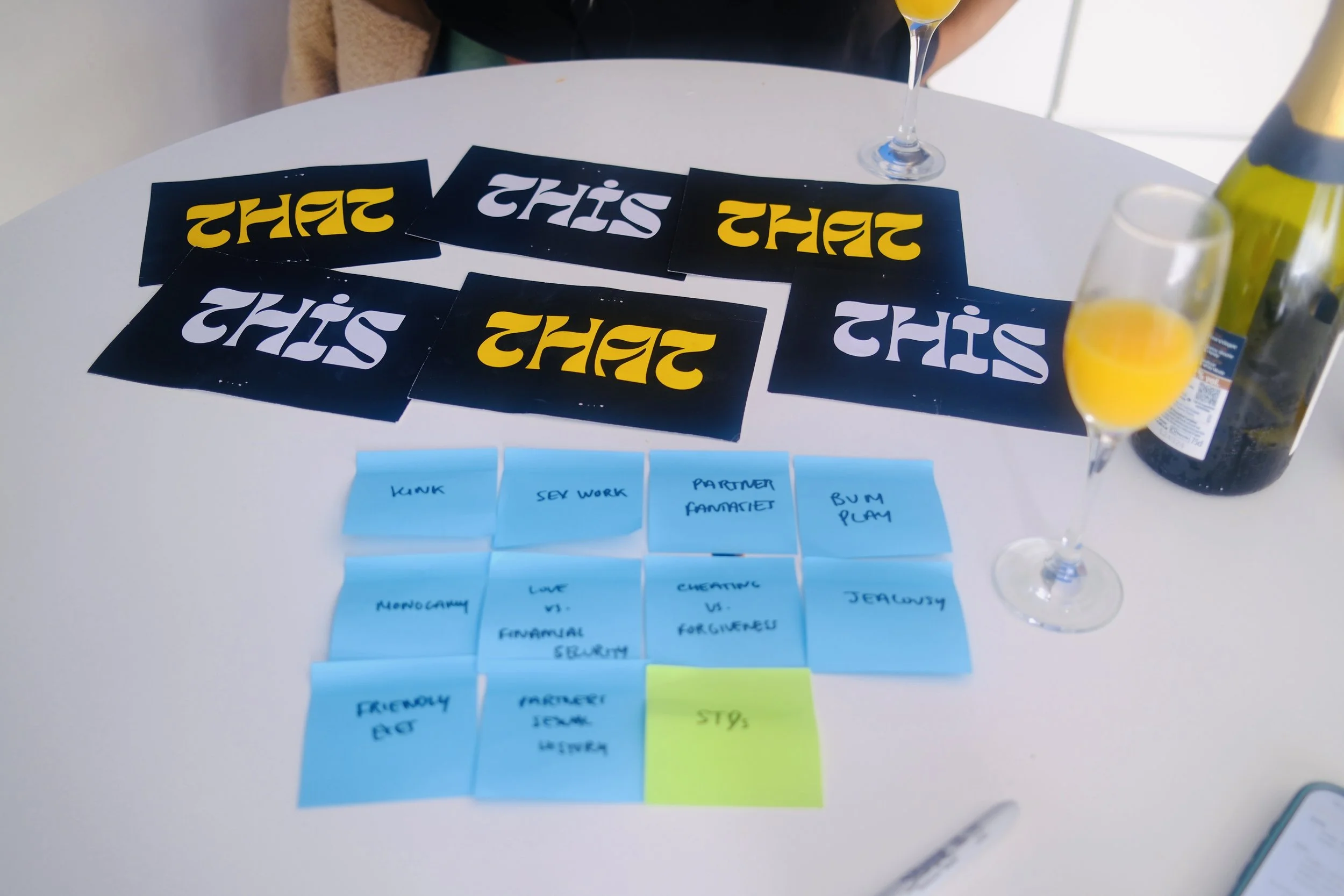 A round table with several blue and black cards, some yellow papers, a pen, a pen, a bottle of wine, and two glasses of orange juice.