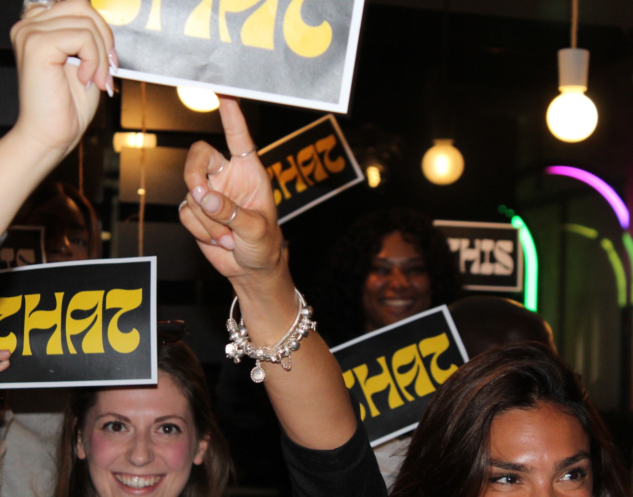 Group of women smiling and holding up signs with the word 'SHIFT' written in yellow and black, in a lively indoor setting with hanging lights and colorful neon lights in the background.