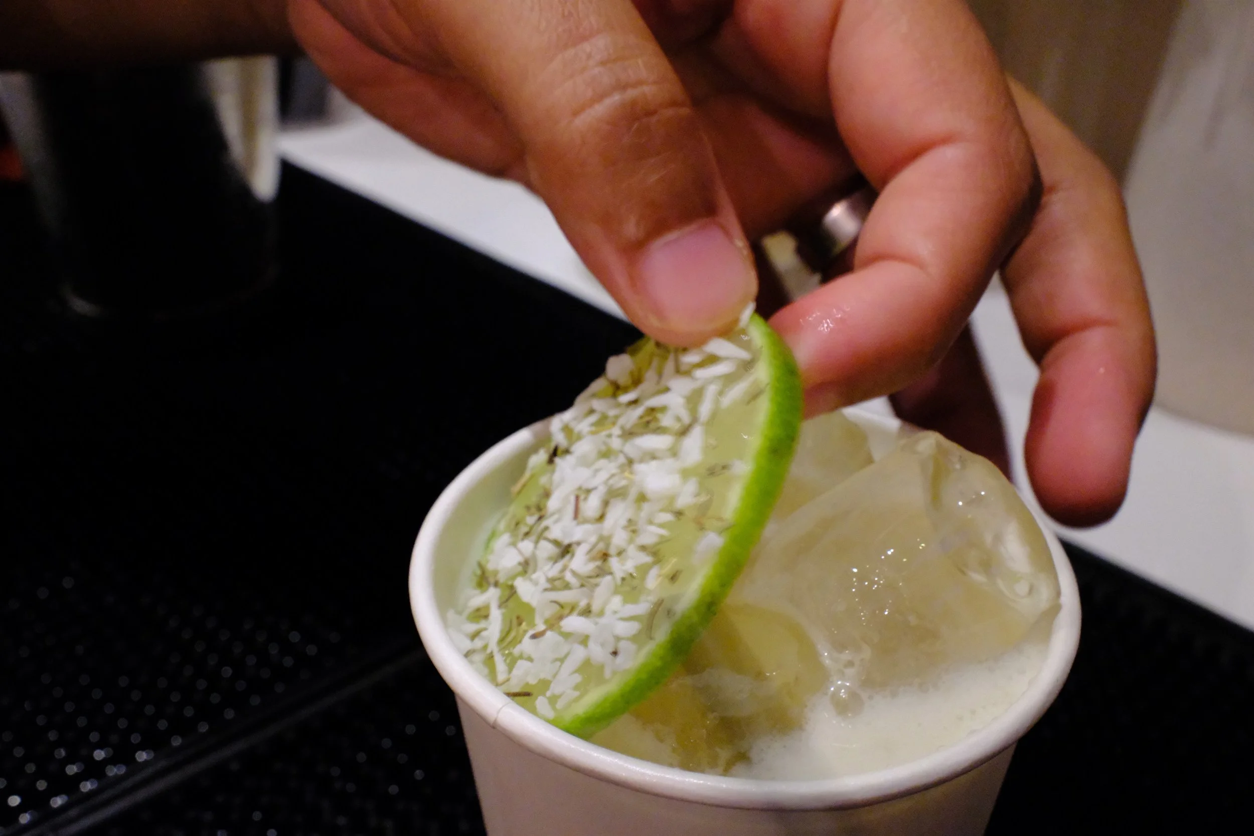 A hand holding a lime wedge garnished with shredded coconut, being placed on top of a drink with ice and creamy light-colored liquid in a paper cup.
