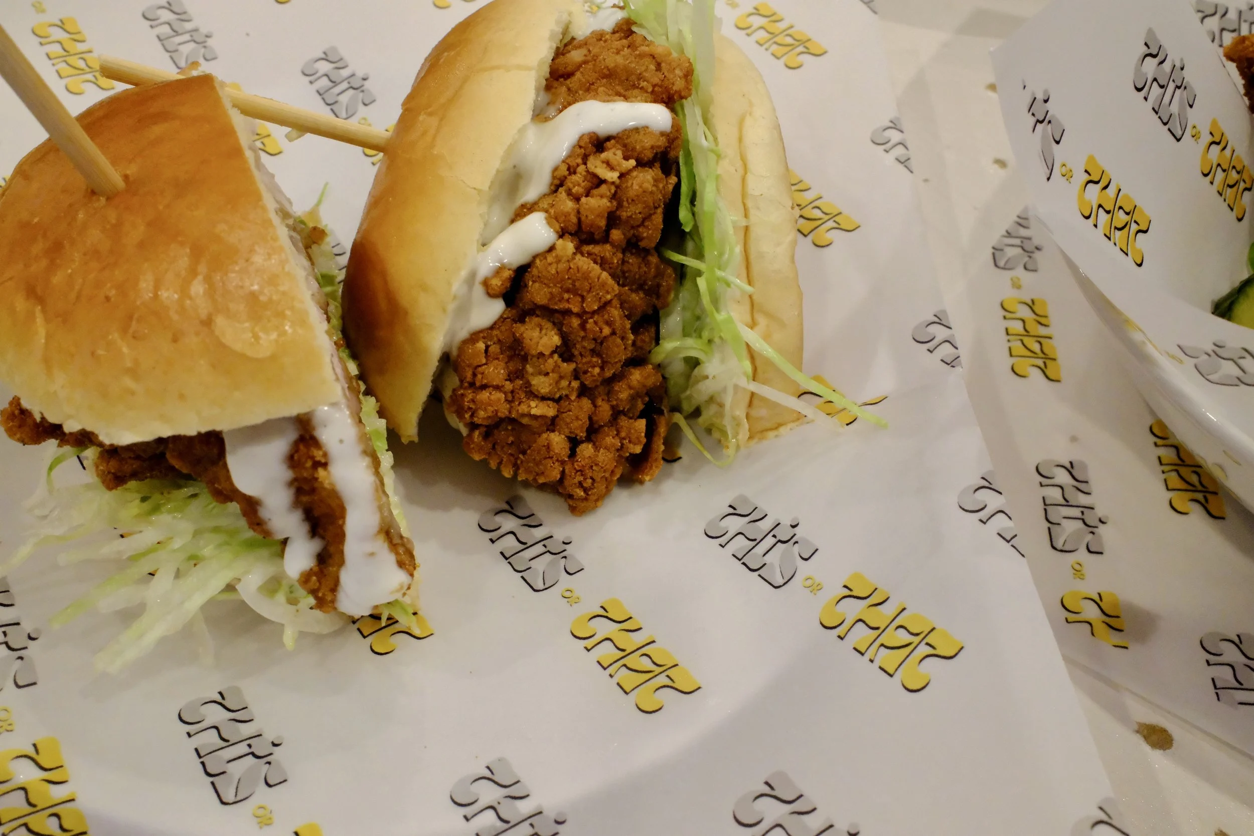 Two fried chicken sandwiches with shredded lettuce, mayonnaise, and toasted buns, served on branded paper with yellow and black text.