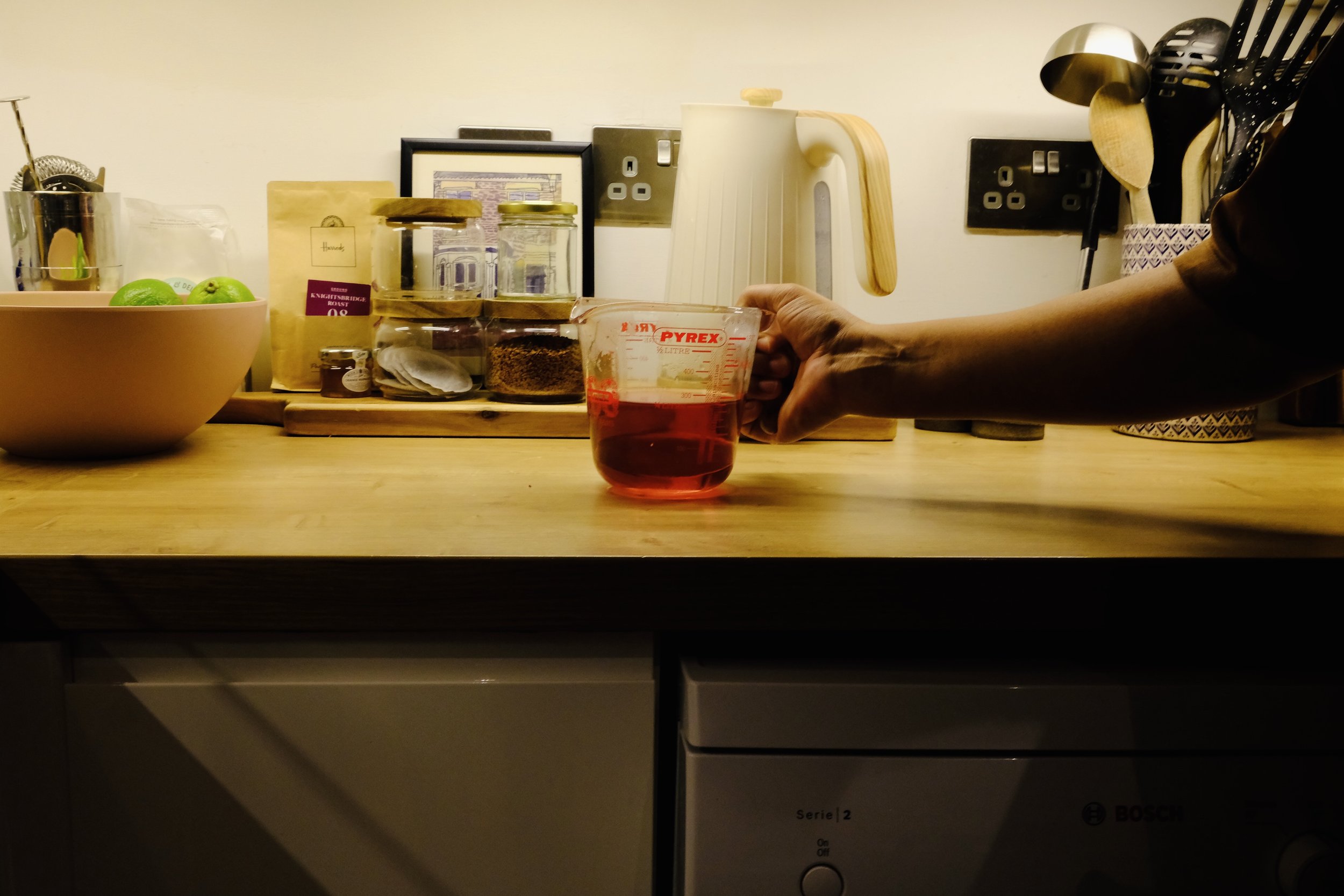 A kitchen countertop with jars, a framed picture, a cream-colored kettle, and a container holding wooden spoons and black utensils. A person's arm is reaching out to grasp a measuring cup filled with red liquid.