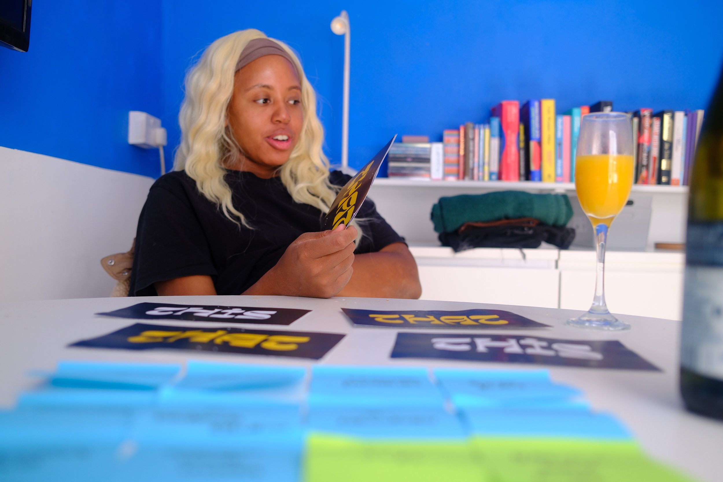 Woman with blonde wavy hair, wearing a black t-shirt, sitting at a table with flyers, holding one flyer in her hand, in a room with blue walls, a bookshelf, and a glass of orange juice on the table.