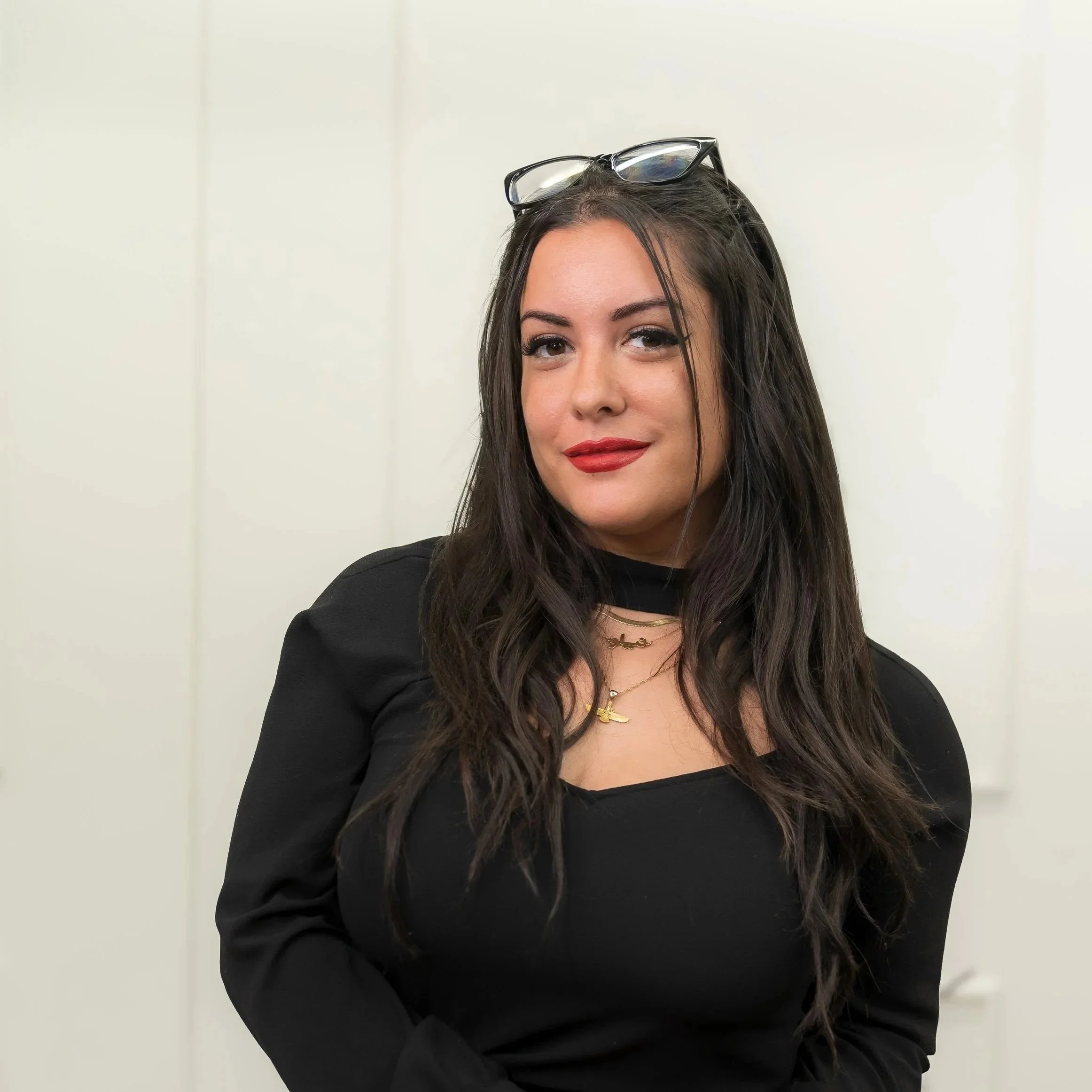 A woman with long, wavy black hair and light skin, wearing red lipstick, black eyeliner, a gold necklace, and a beige and black patterned top, looking at the camera with a slight smile against a plain grey background.