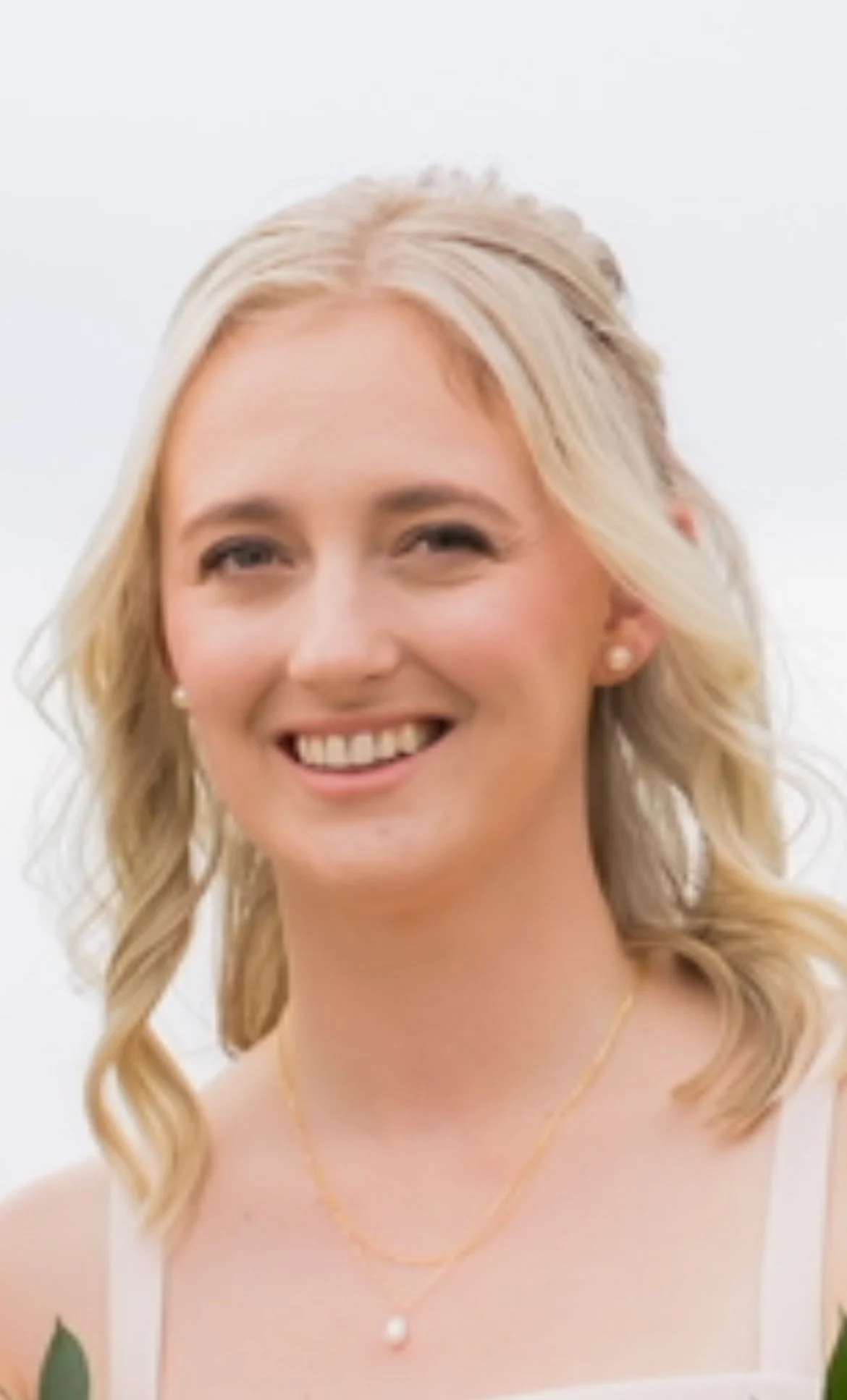 Close-up of a smiling woman with blonde curly hair, wearing pearl earrings and a necklace, with a bright background.