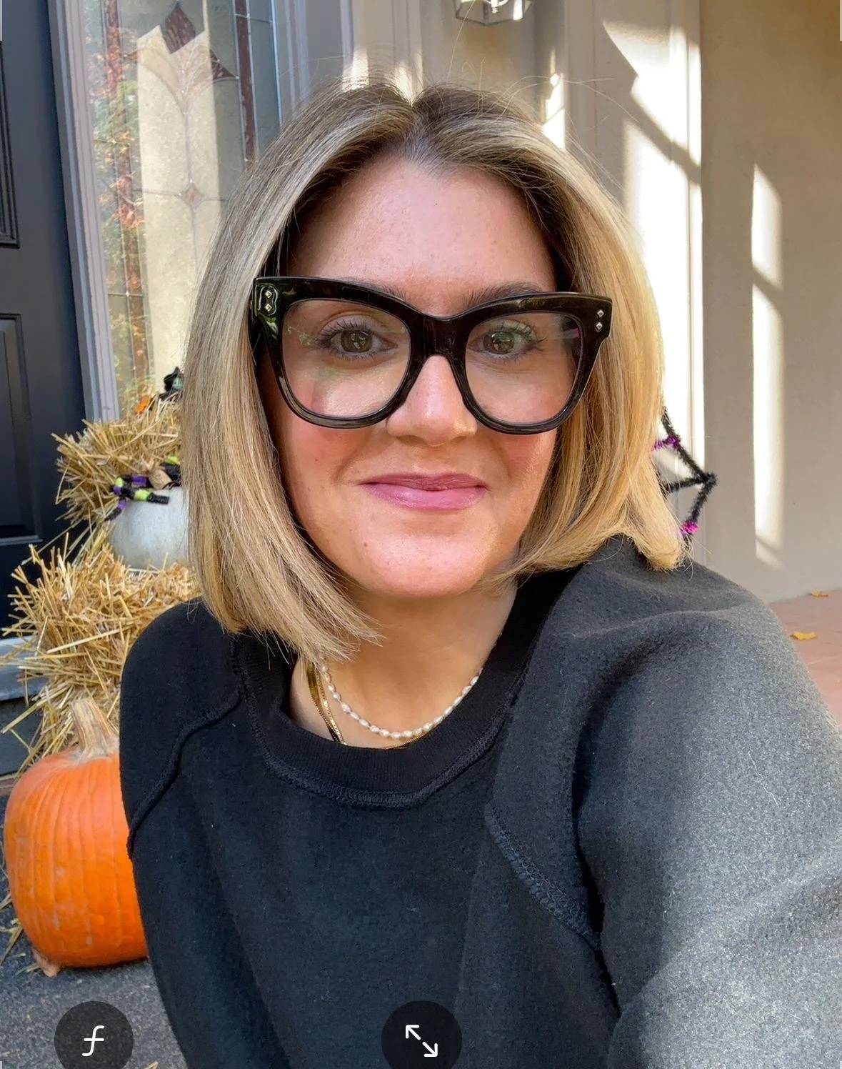A woman with shoulder-length blonde hair and glasses taking a selfie outside during fall, with pumpkins and hay bales decorated for the season visible in the background.