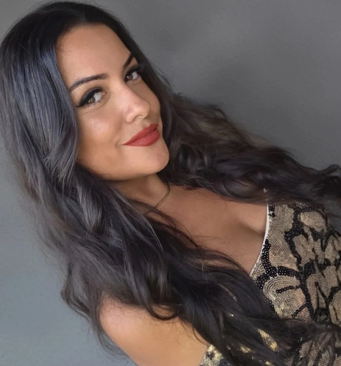 A woman with long, wavy black hair and light skin, wearing red lipstick, black eyeliner, a gold necklace, and a beige and black patterned top, looking at the camera with a slight smile against a plain grey background.
