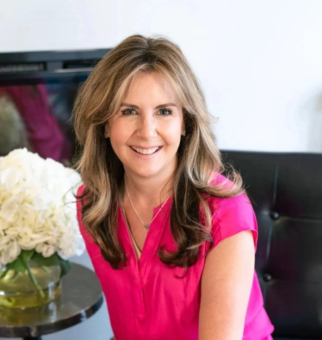 A smiling woman with wavy brown hair wearing a pink blouse and blue jeans, sitting on a black chair next to a table with a vase of white hydrangeas.