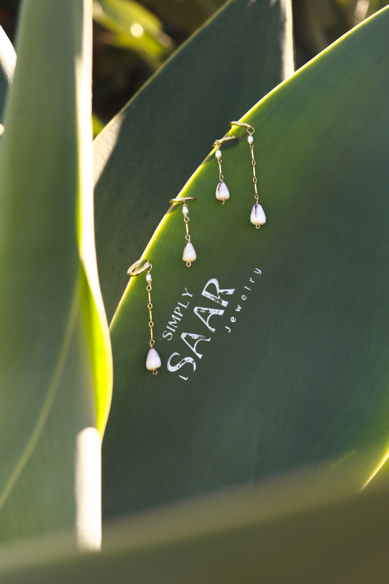 A pair of gold chain earrings with white teardrop-shaped pearls hanging from it, placed over a dark green paper with the logo 'Simply Island Jewelry' printed on it, surrounded by green plant leaves.