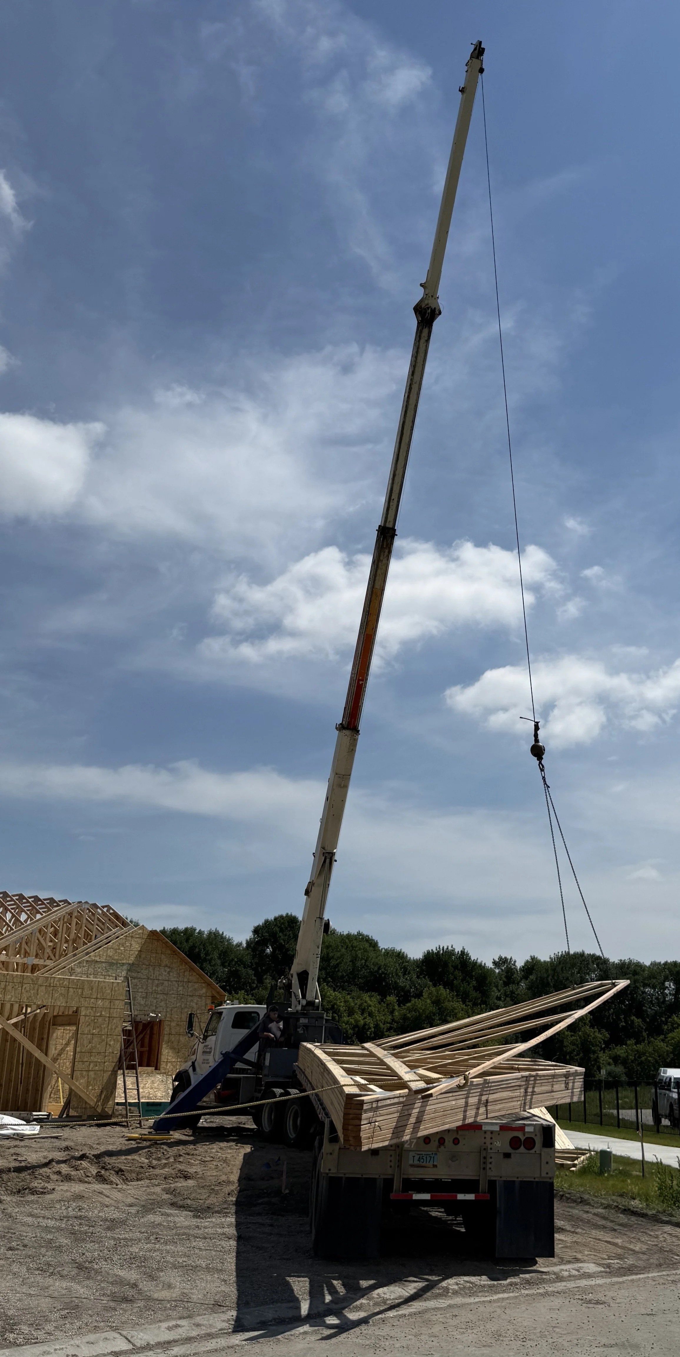 A construction crane lifting wooden roof trusses at a house construction site under a partly cloudy sky.