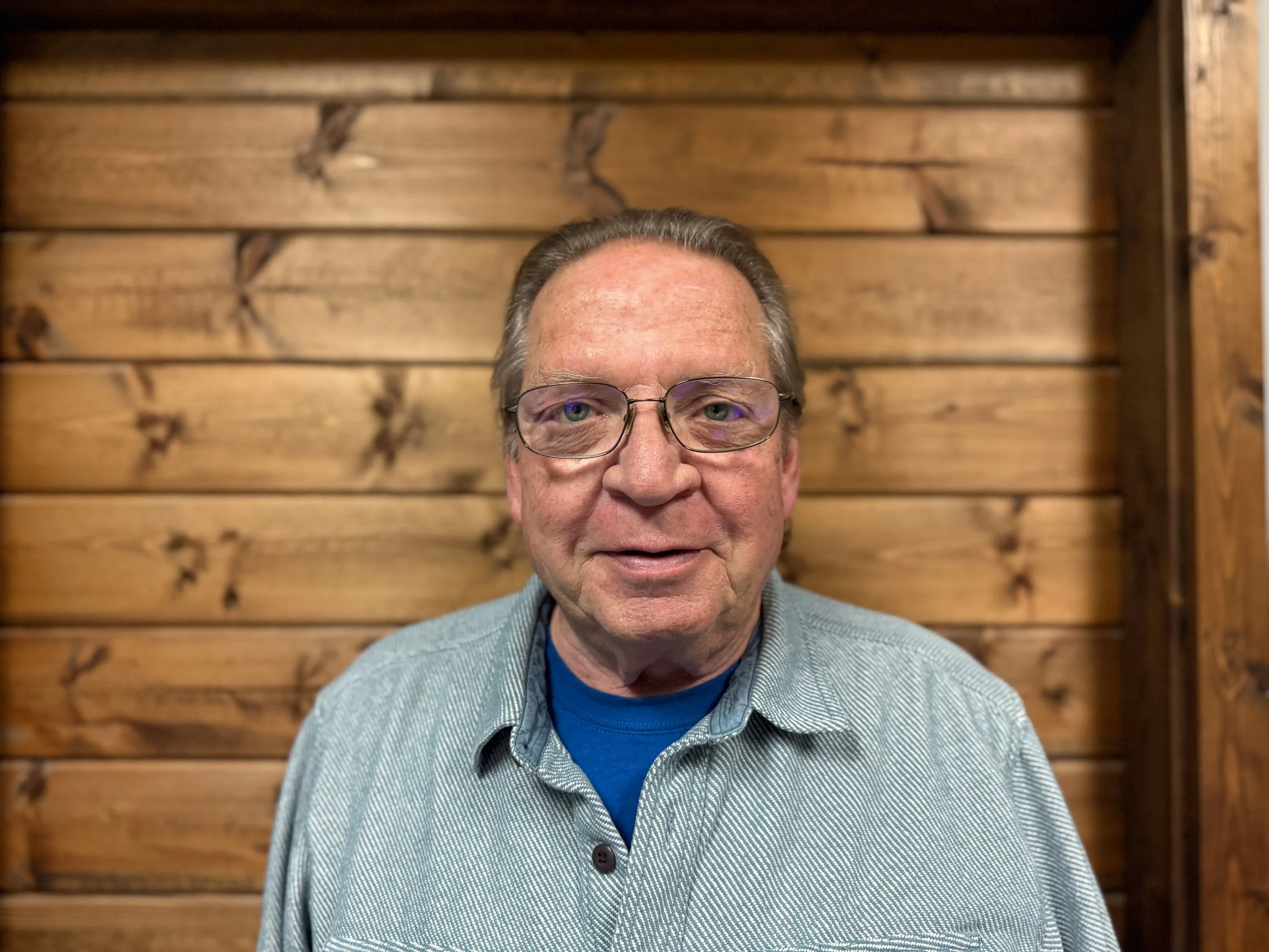 A close-up portrait of an older man with glasses, wearing a blue shirt and gray jacket, standing in front of a wooden wall.