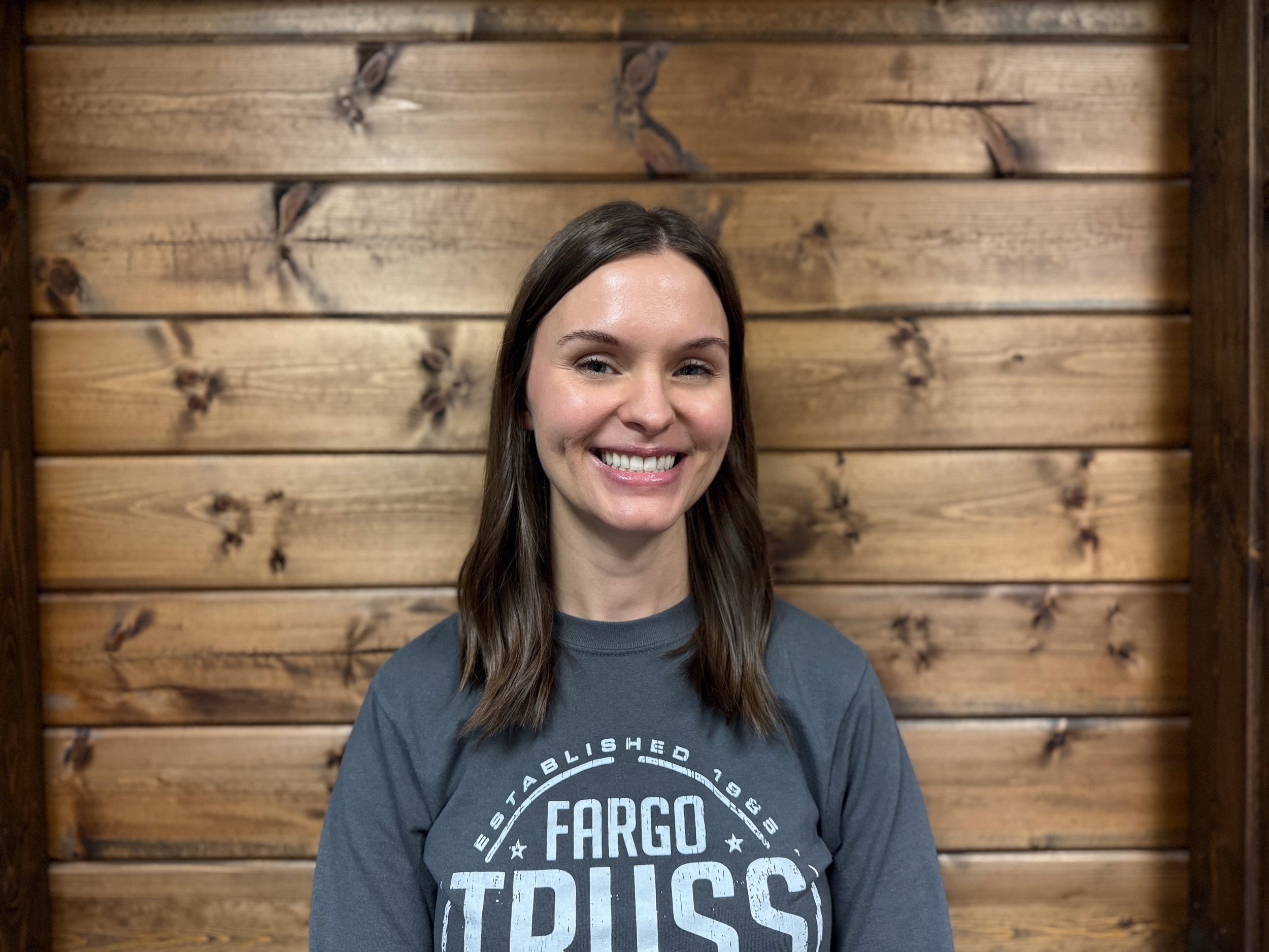 A woman with long brown hair smiling in front of a wood-paneled wall, wearing a gray shirt with white text that says Fargo Truss