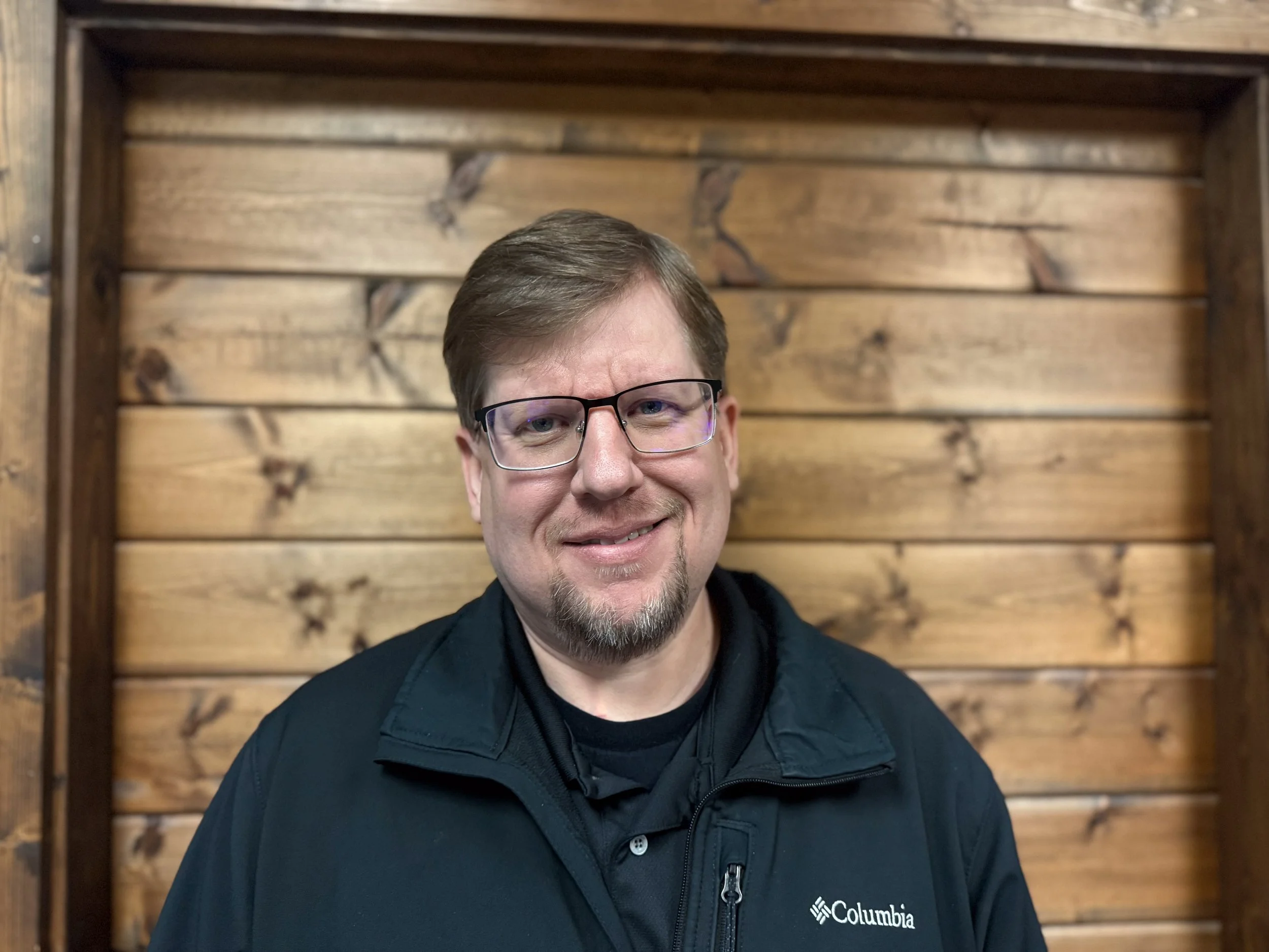 A man with glasses, brown hair, and a goatee, smiling while wearing a black Columbia jacket, standing in front of a wooden wall.