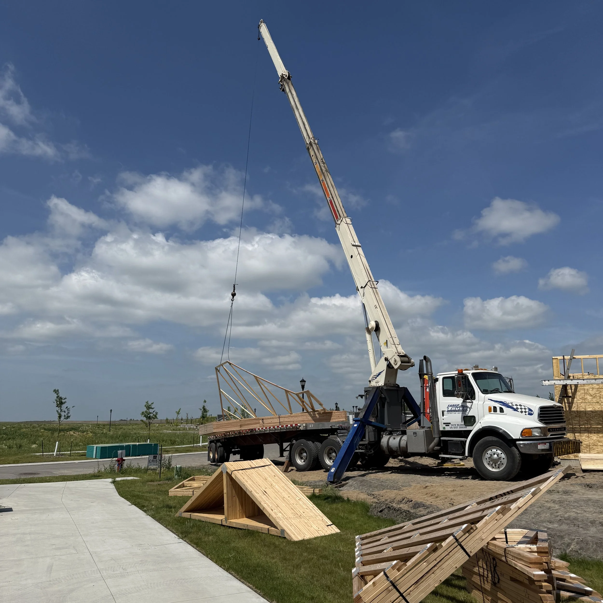A construction site with a crane lifting wooden framing components, with construction materials and partially built structures nearby, under a partly cloudy sky.