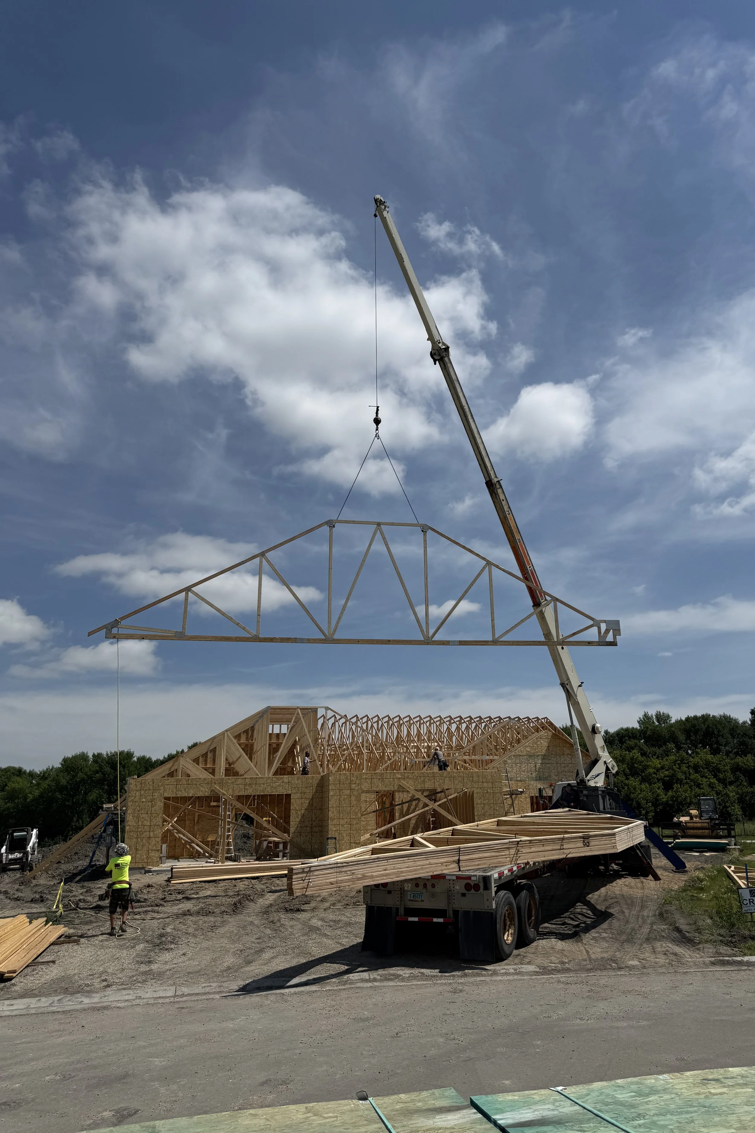 Construction site with wooden framing and a crane lifting a large truss piece into place under a partly cloudy sky.