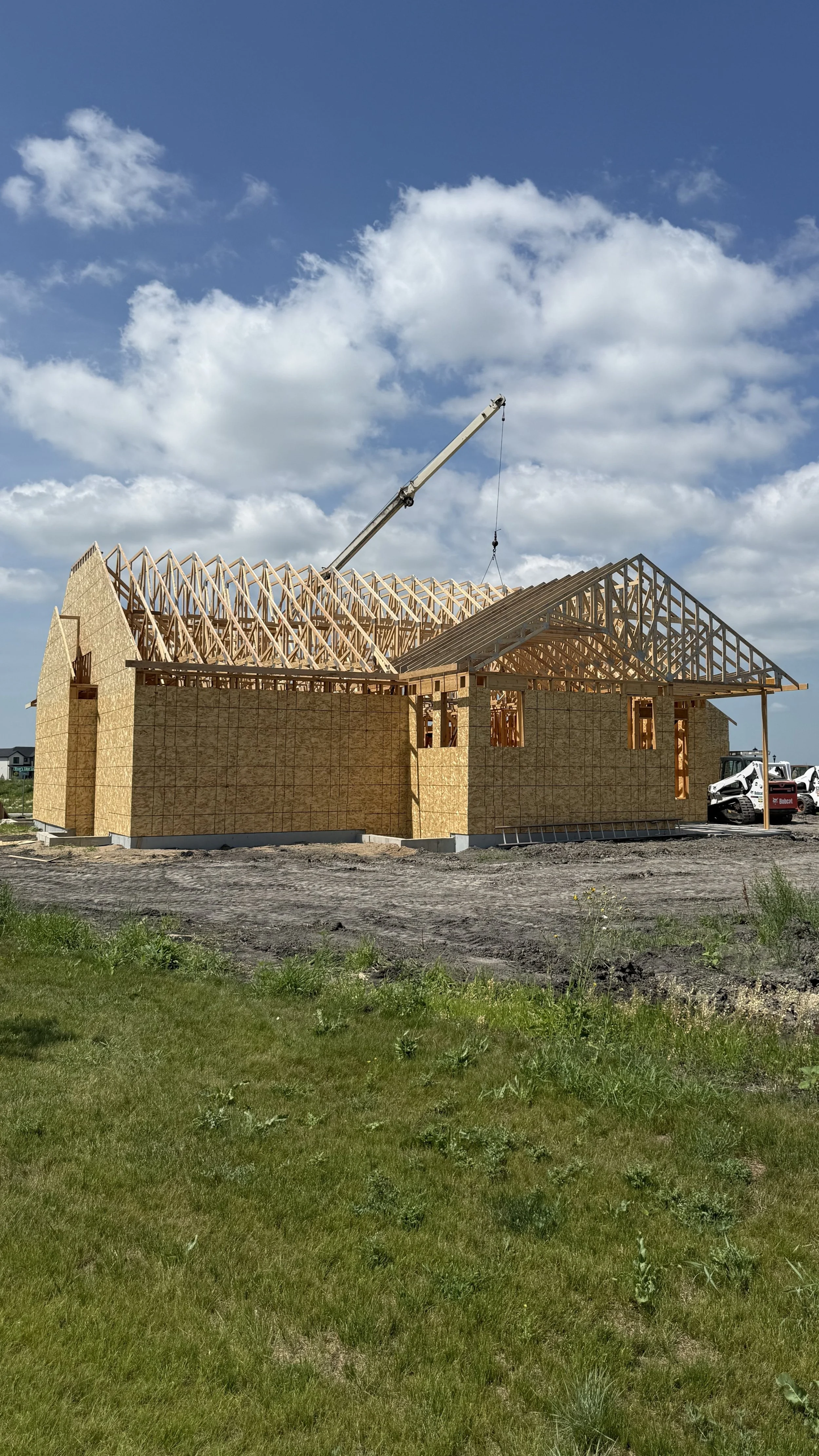 A house under construction with wooden framing and a crane in the background, set against a blue sky with scattered clouds.
