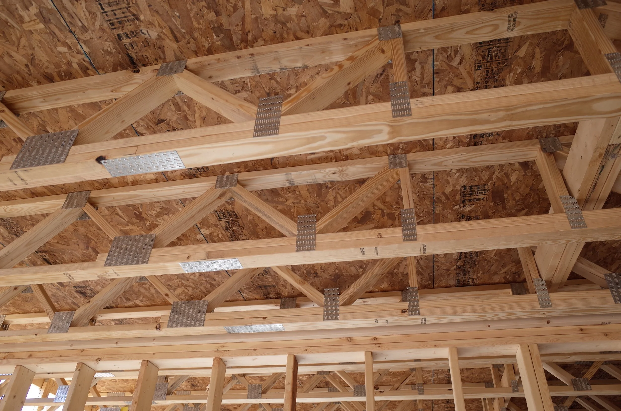 Interior view of building framing showing wooden trusses and OSB sheathing, with metal connectors and nails.