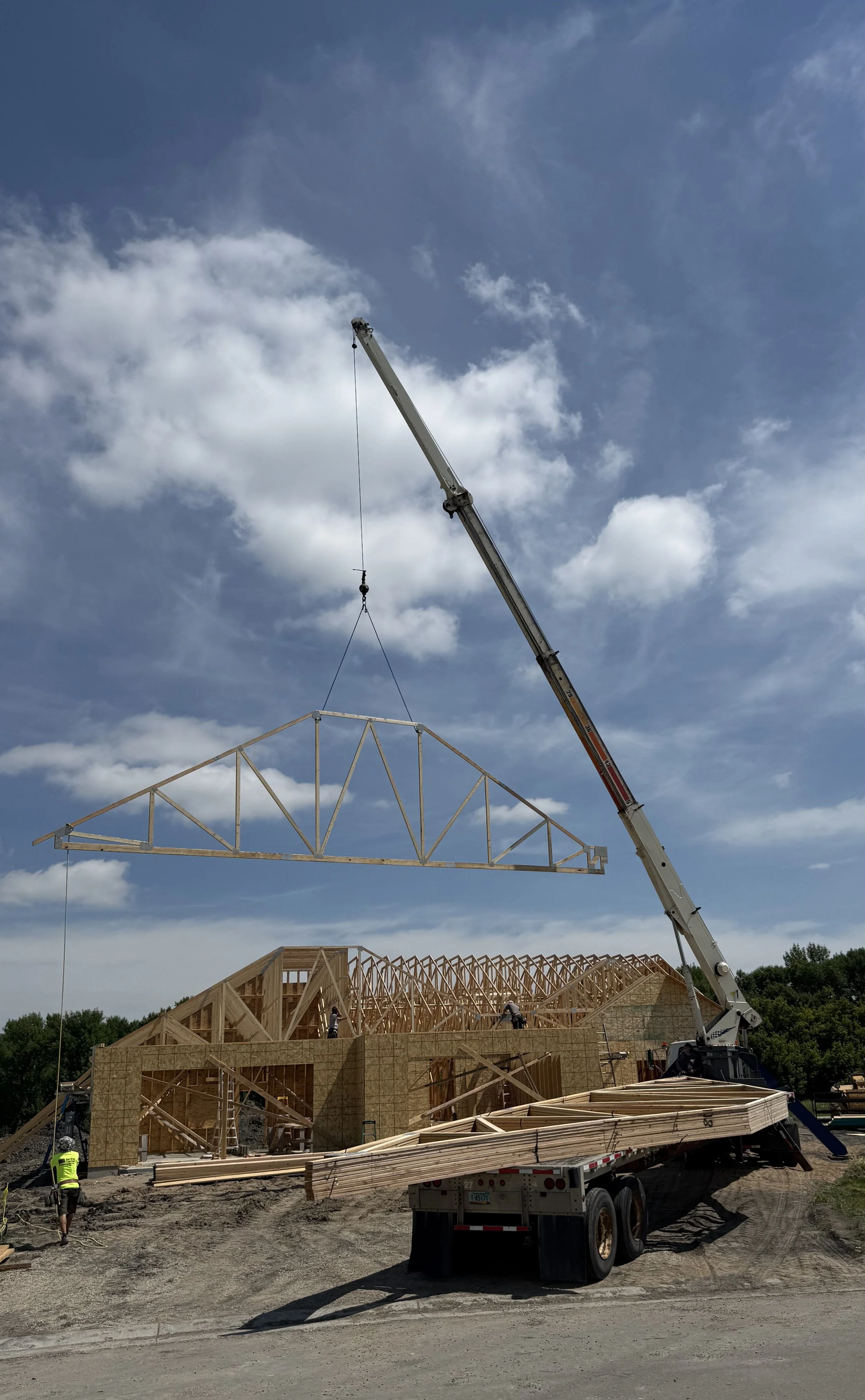 Construction site with wooden framing for a building, crane lifting a truss, workers on site, and blue sky with scattered clouds.